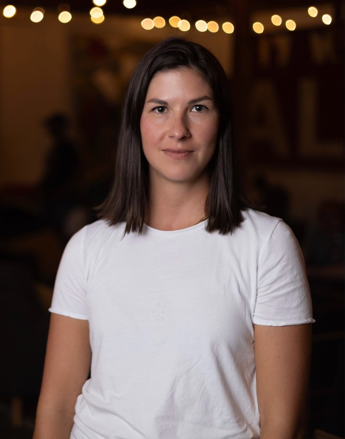 Hego Portrait of a woman with shoulder-length dark hair, wearing a white t-shirt, in a warmly lit indoor setting with string lights in the background.