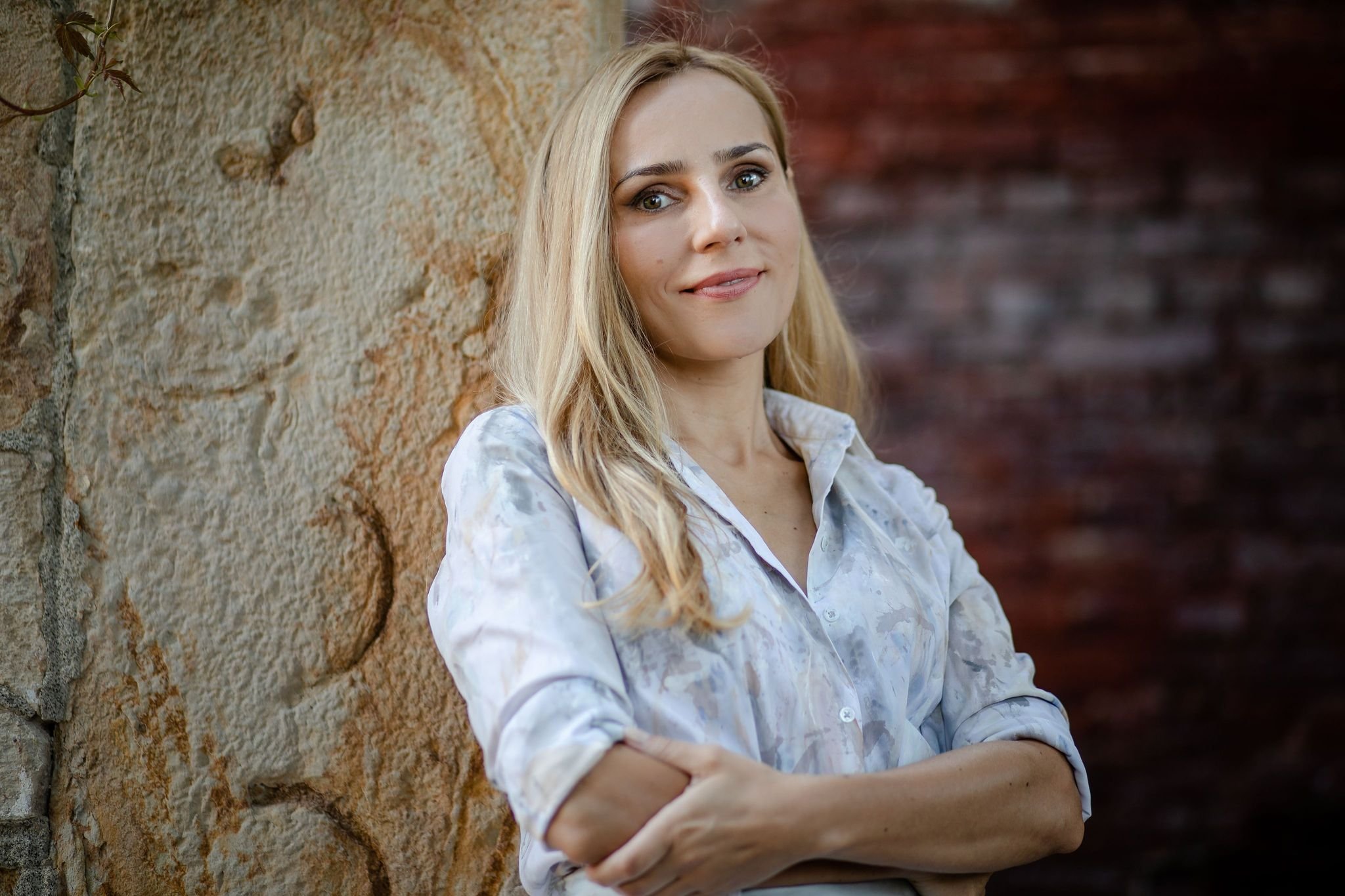 Zana Ranđelović Brown A woman with blonde hair standing with her arms crossed, leaning against a rustic stone wall, and looking at the camera.