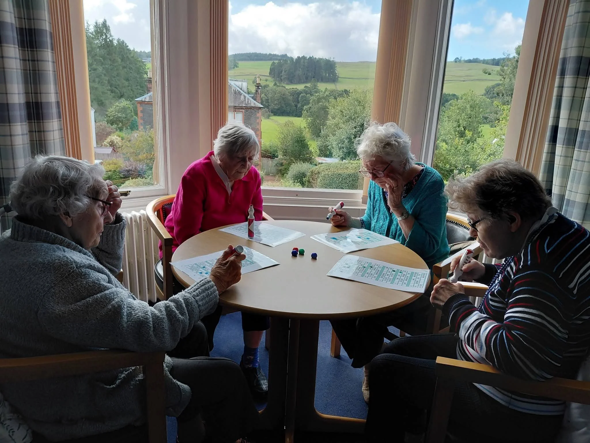 Five elderly women seated around a table playing bingo, with bingo cards and small colorful balls, near large windows overlooking a green countryside landscape.