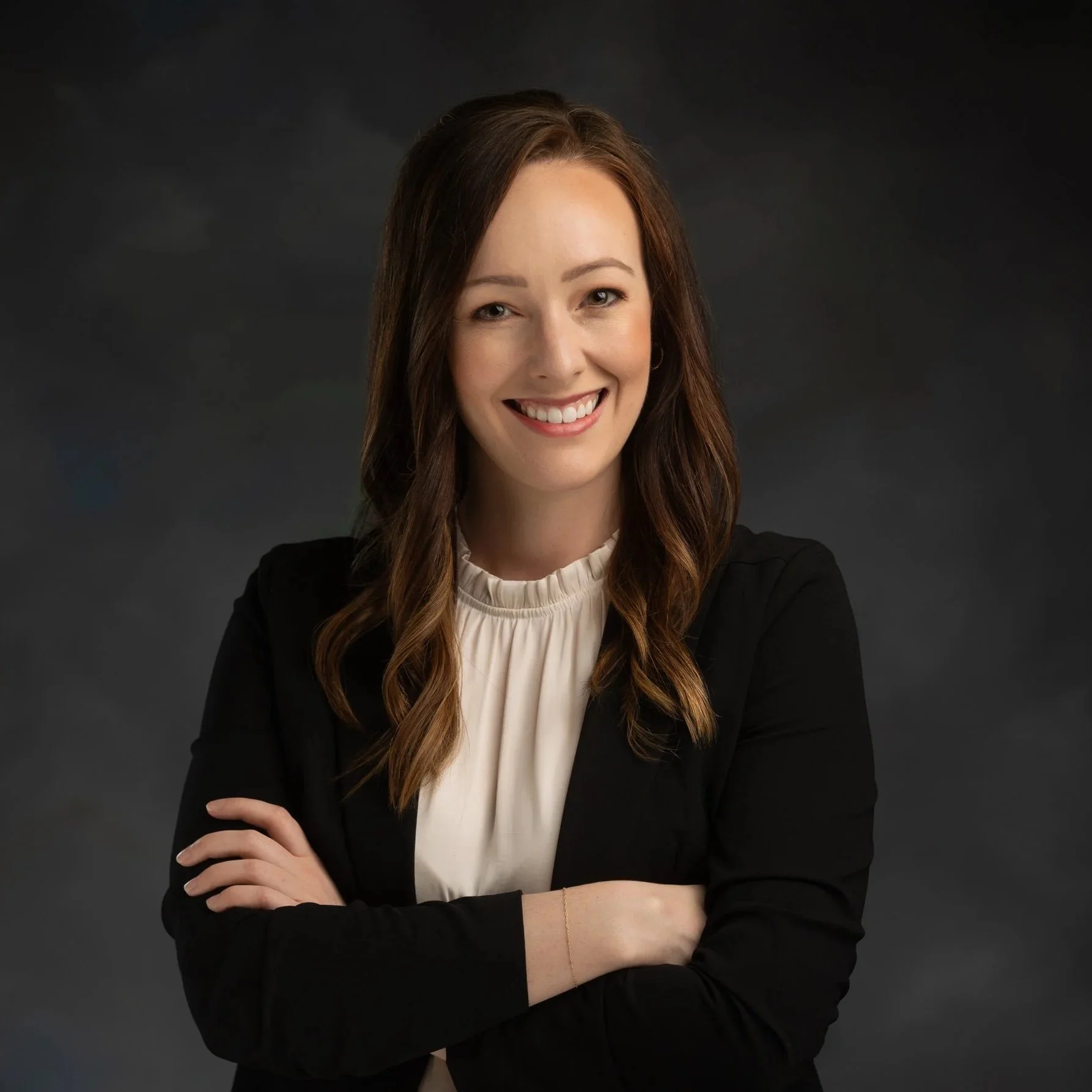 Portrait of a smiling woman with long brown hair, wearing a black blazer and a cream blouse, crossing her arms, against a dark gray background.