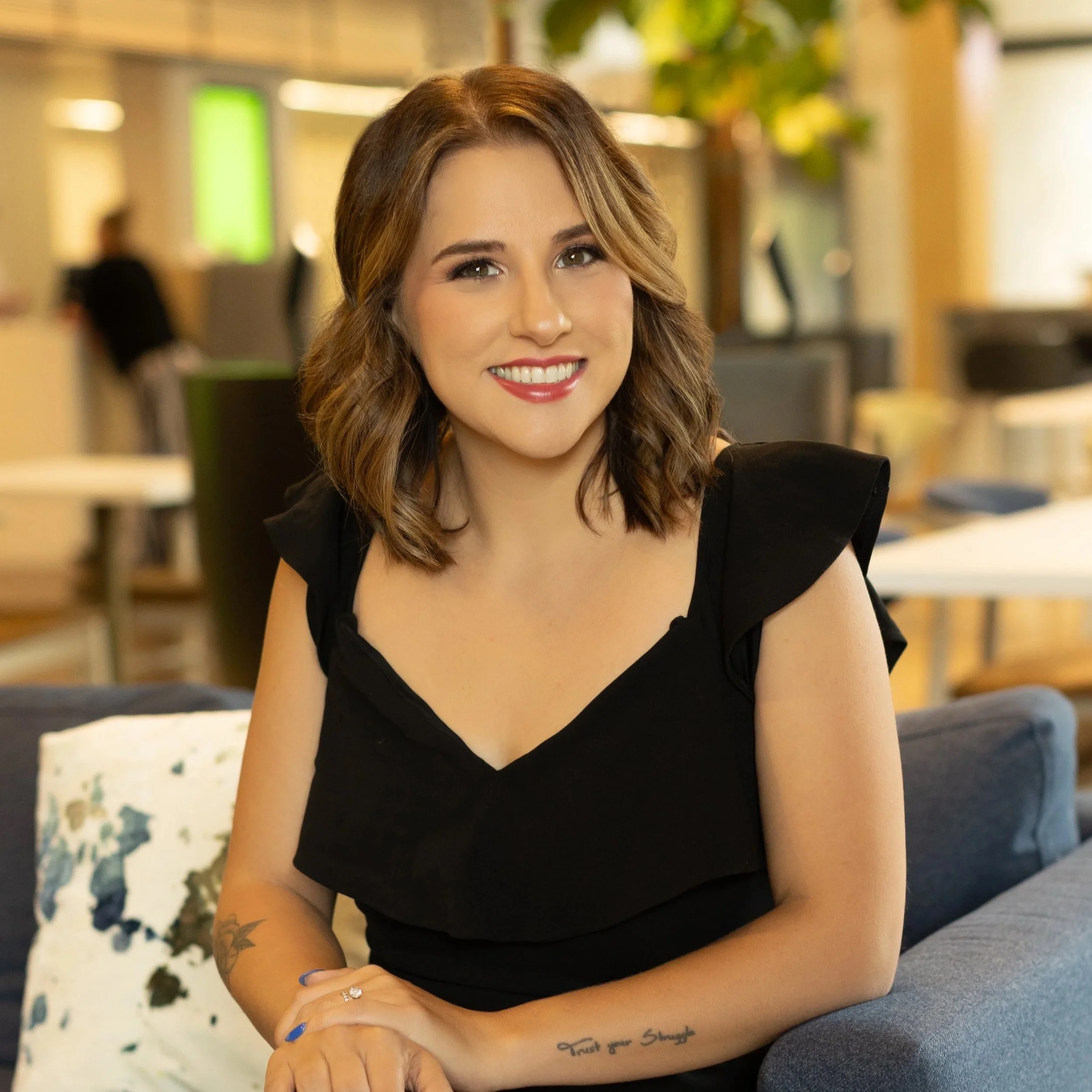 A woman with brown, wavy hair wearing a black top, sitting on a couch in a brightly lit indoor space.