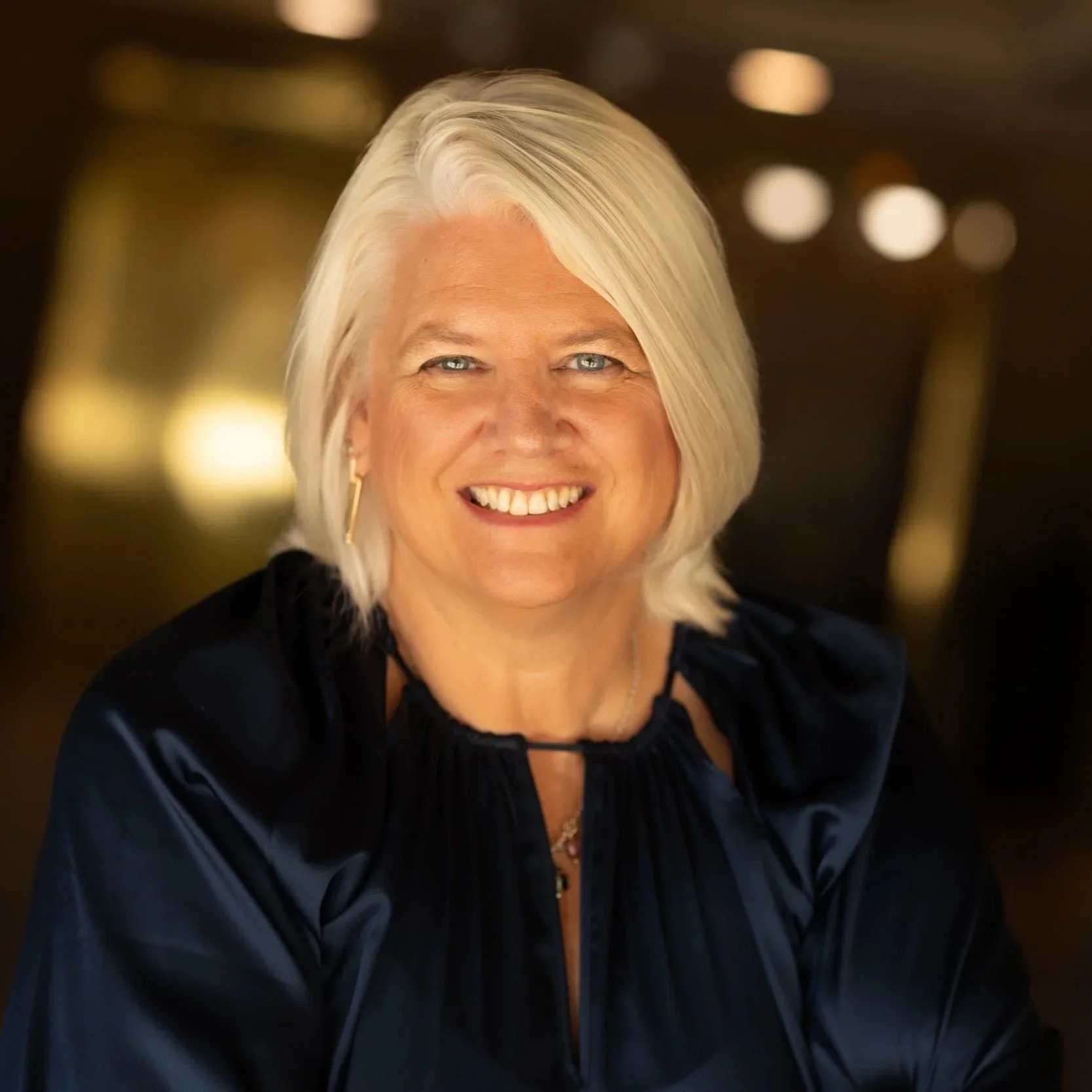 A middle-aged woman with blonde hair and blue eyes, smiling, wearing a dark blue satin blouse with a decorative collar and hoop earrings, in a warmly lit indoor setting.