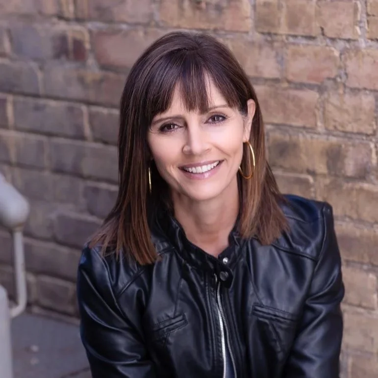 A woman with shoulder-length brown hair, wearing gold hoop earrings and a black leather jacket, smiling in front of a brick wall.