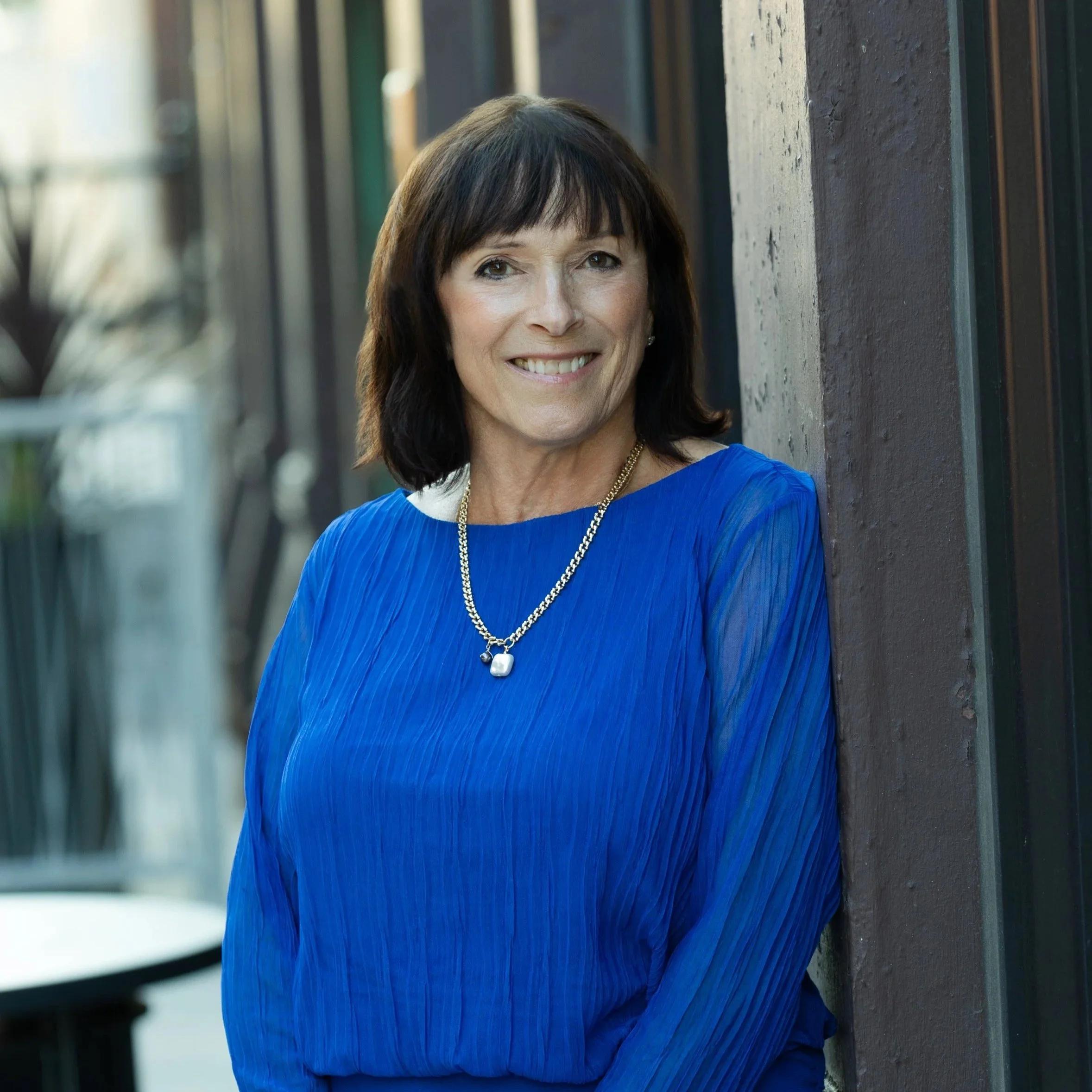 A woman with dark brown hair and a bright smile, wearing a vibrant blue dress with sheer sleeves and a gold necklace, standing outdoors against a dark wall with blurred background.