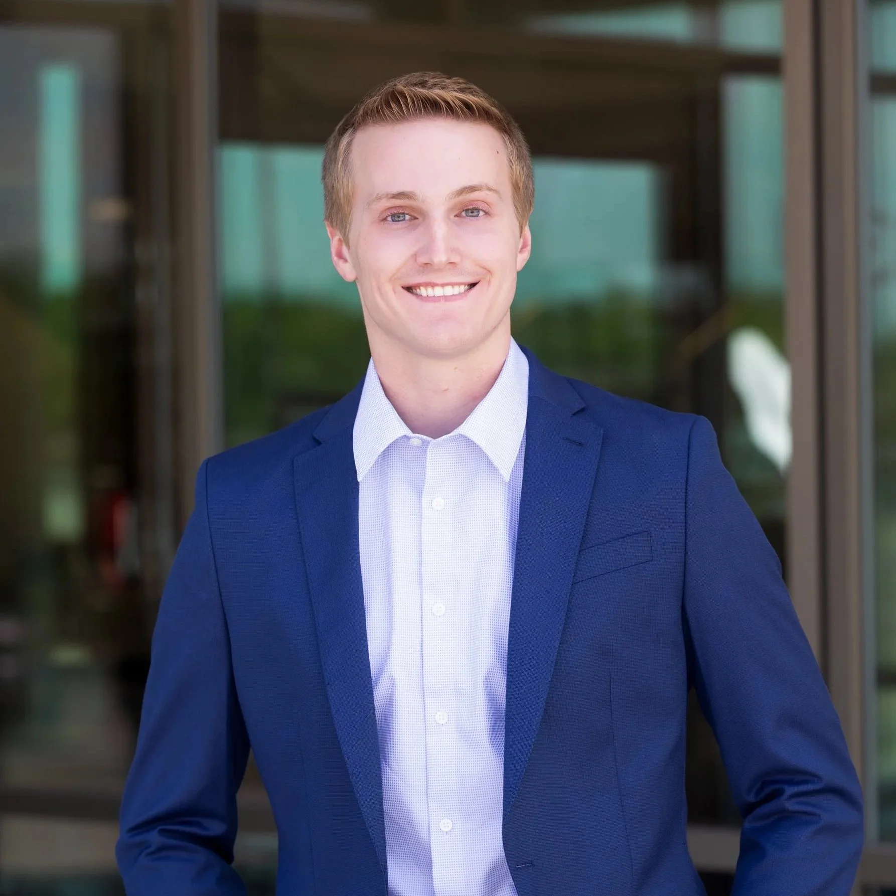 A young man in a blue suit and white dress shirt standing outdoors in front of a modern building with glass windows, smiling at the camera.