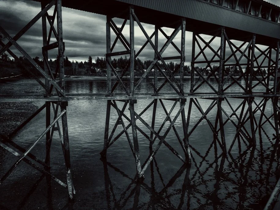 black and white image of underneath a bridge with the reflection of the post in the water
