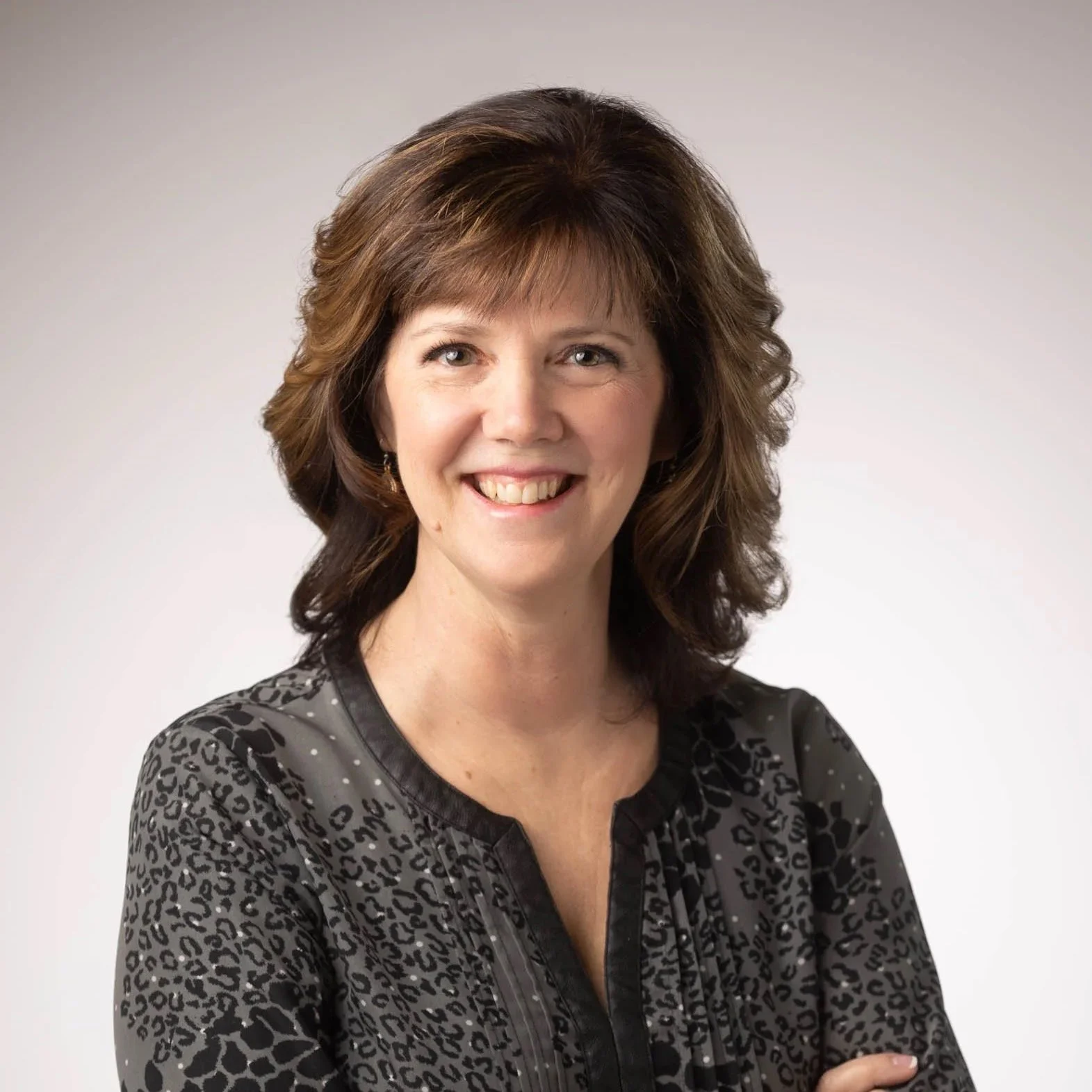 A smiling woman with short, wavy brown hair wearing a patterned dark blouse, posing against a plain light background.