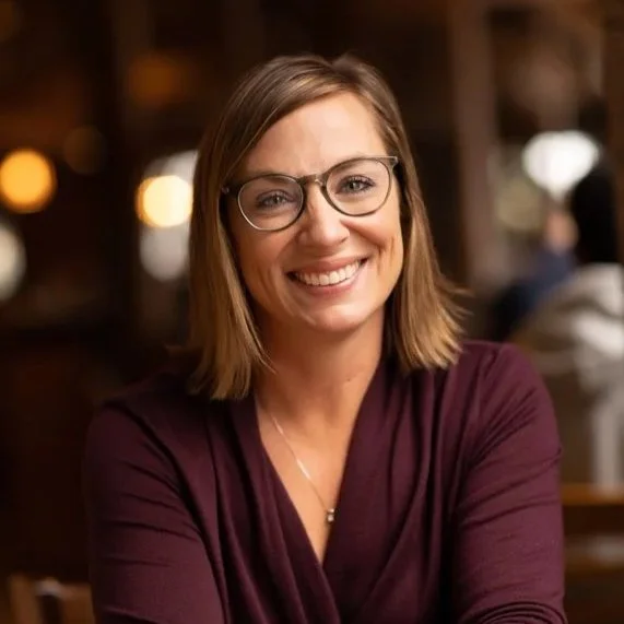 A smiling woman with shoulder-length brown hair, glasses, and a maroon top, sitting in a warmly lit indoor environment.