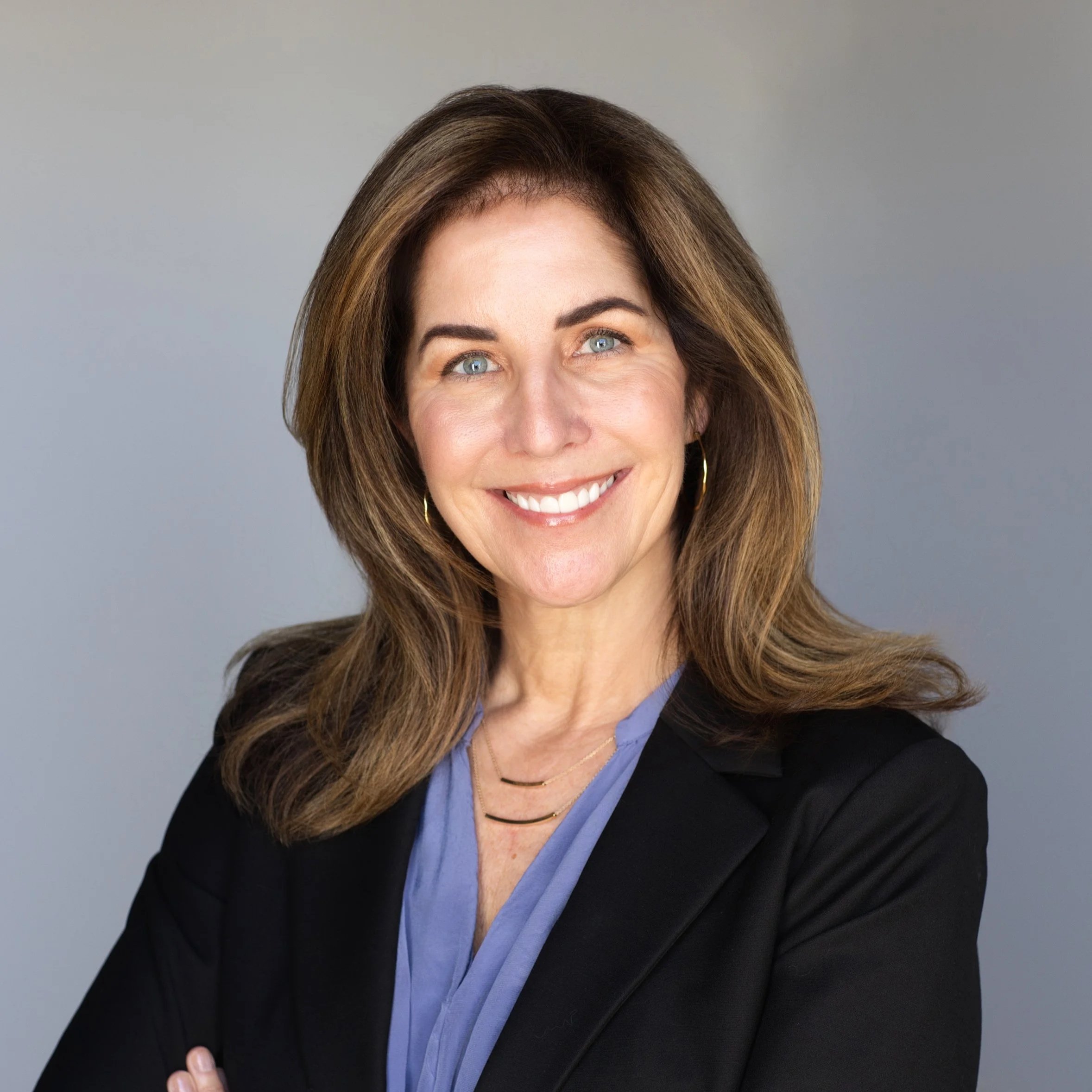 A professional woman with shoulder-length brown hair, blue eyes, and a bright smile, wearing a black blazer, a light blue blouse, gold hoop earrings, and a layered gold necklace, standing with arms crossed against a plain gray background.