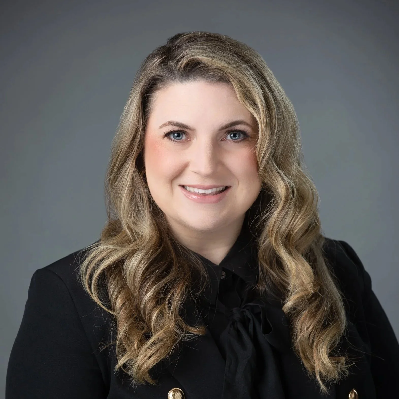 Professional headshot of a woman with long, wavy blonde hair, blue eyes, wearing a black blazer and a black blouse with a bow tie, smiling against a gray background.