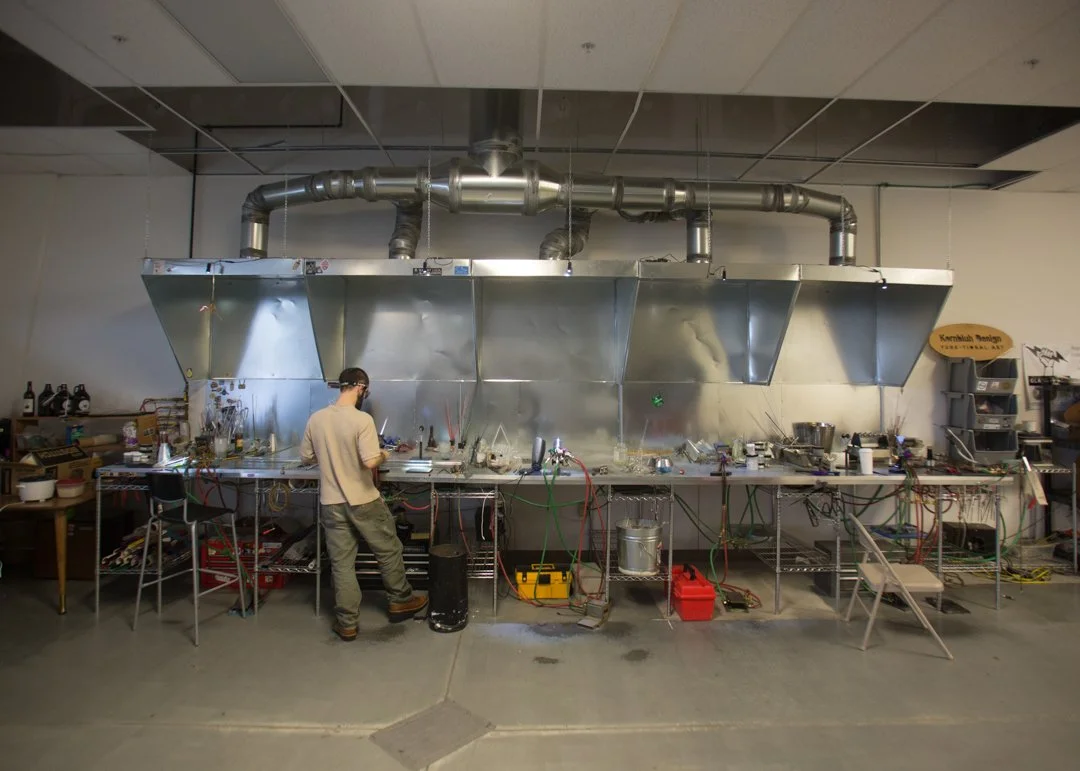 wide image of glass blower at his station with vents at work space in background, shot by commercial photographer Corey Rourke

