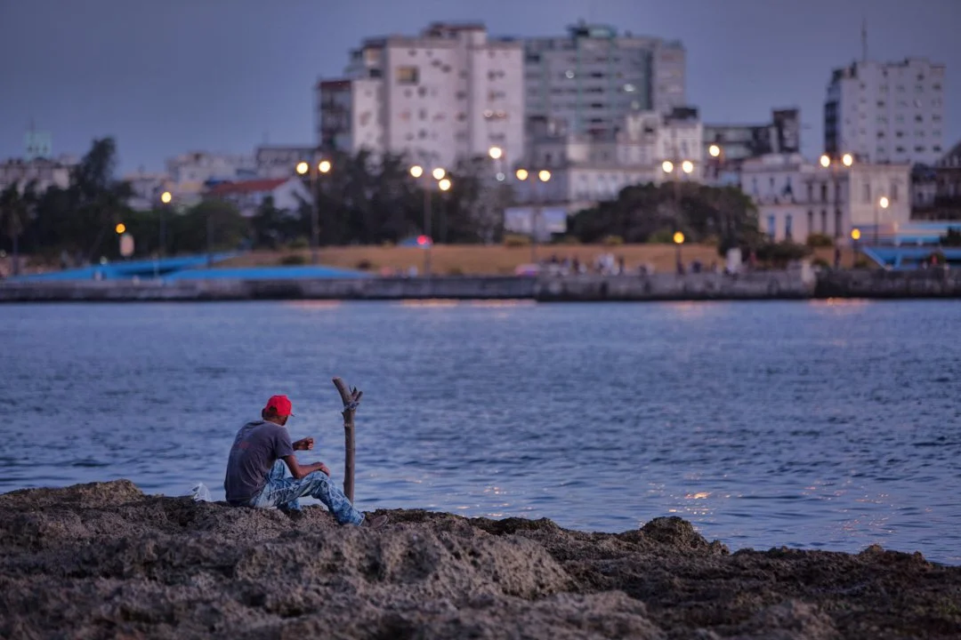 man in red hat fishing with string on the rock coast middle ground water foreground is the city scape in Cuba.  