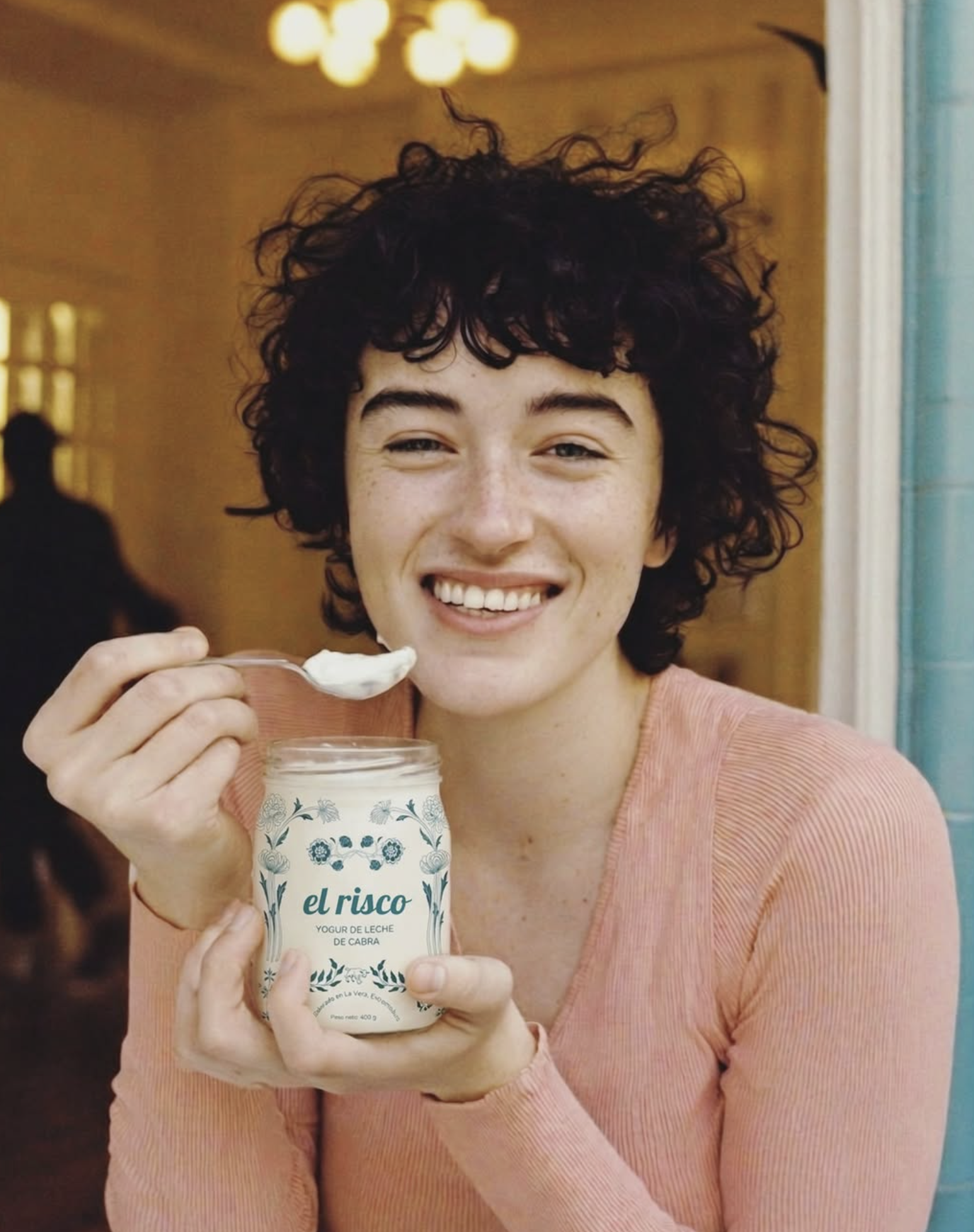 Joven con cabello rizado, sonriendo y sosteniendo un envase de yogur de leche de cabra llamado "el risco" en un lugar bien iluminado.