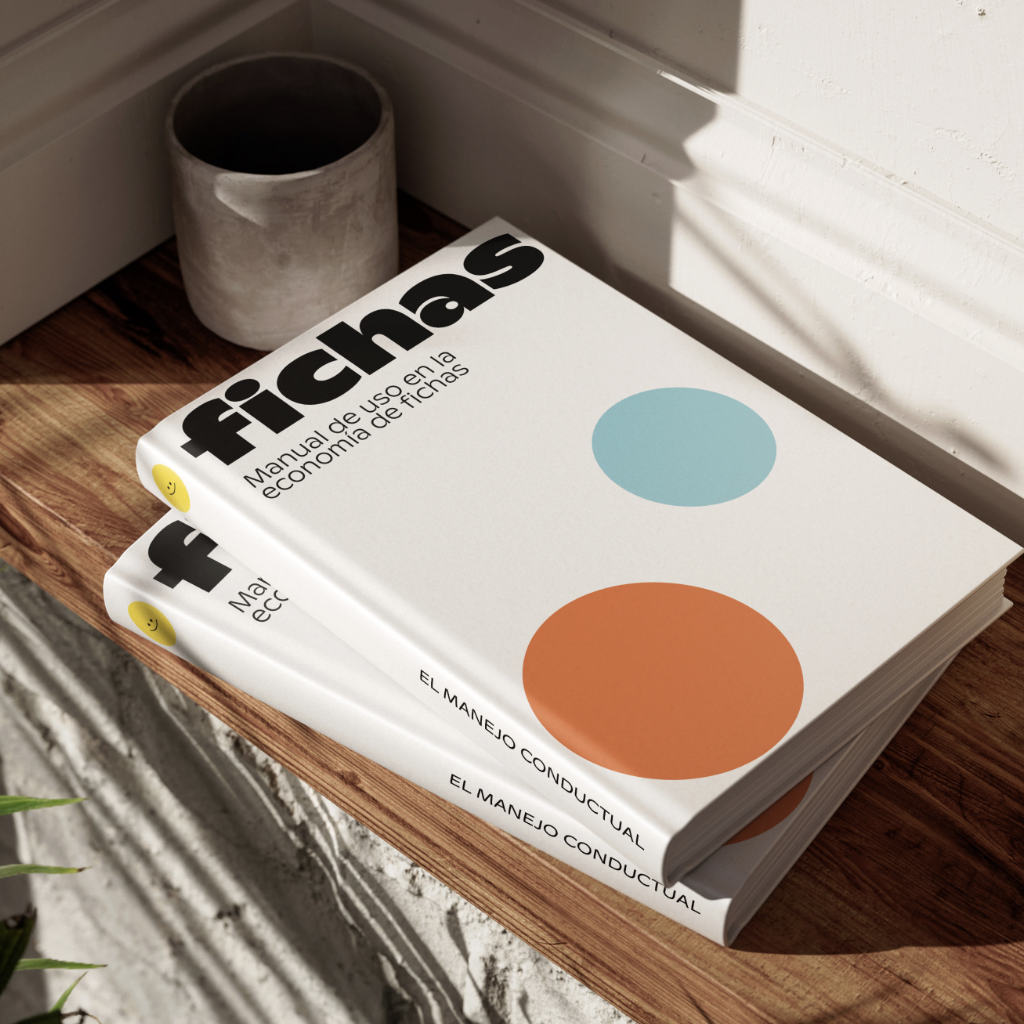 Dos libros apilados titulados "fichas: Manual de uso en la economía de fichas" en una mesa de madera con una maceta gris y una planta verde a la izquierda, en una pared blanca.