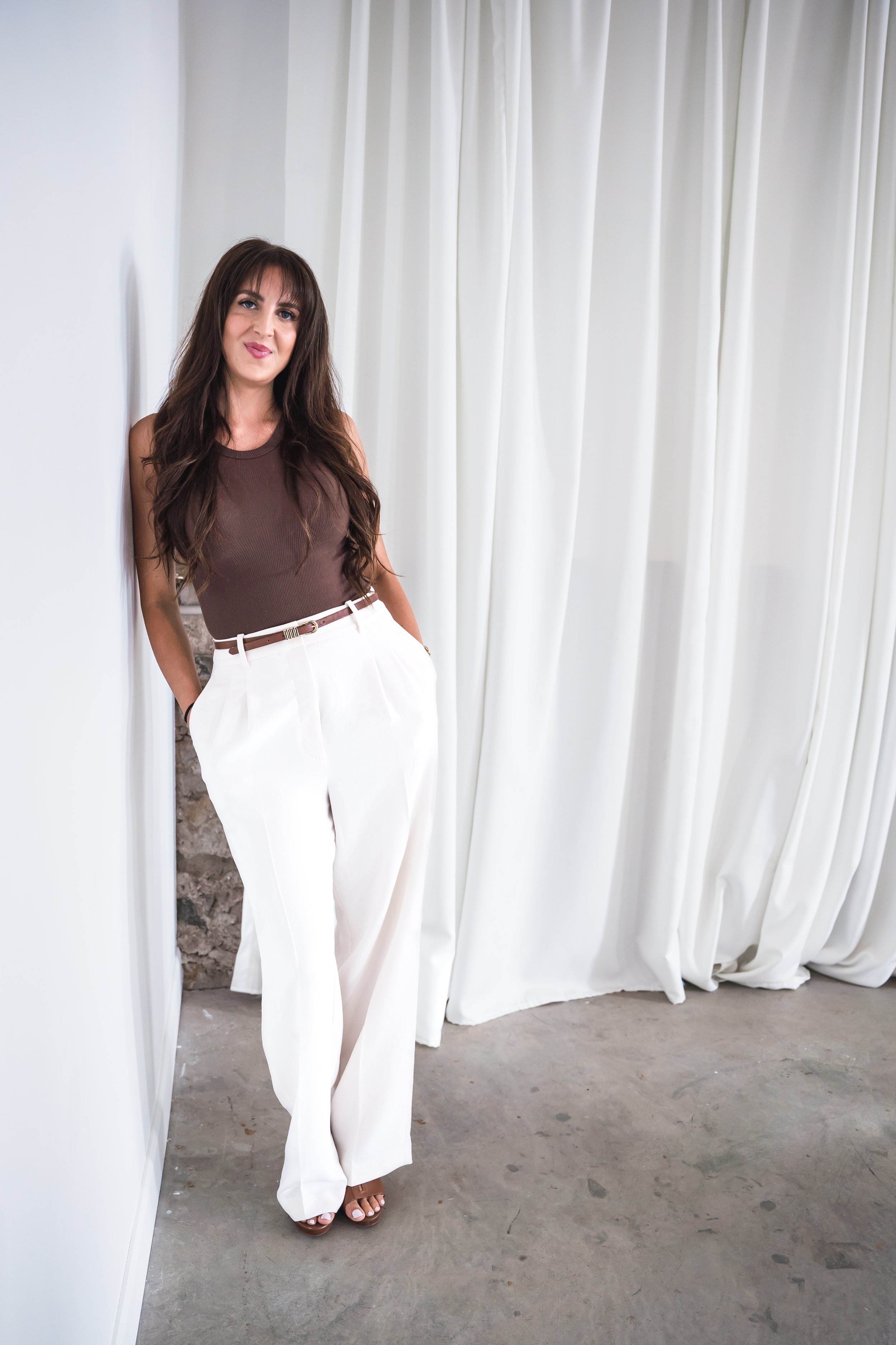 A medical aesthetician in Brantford Ontario leaning on a white wall with her hands in the pockets of her trousers.
