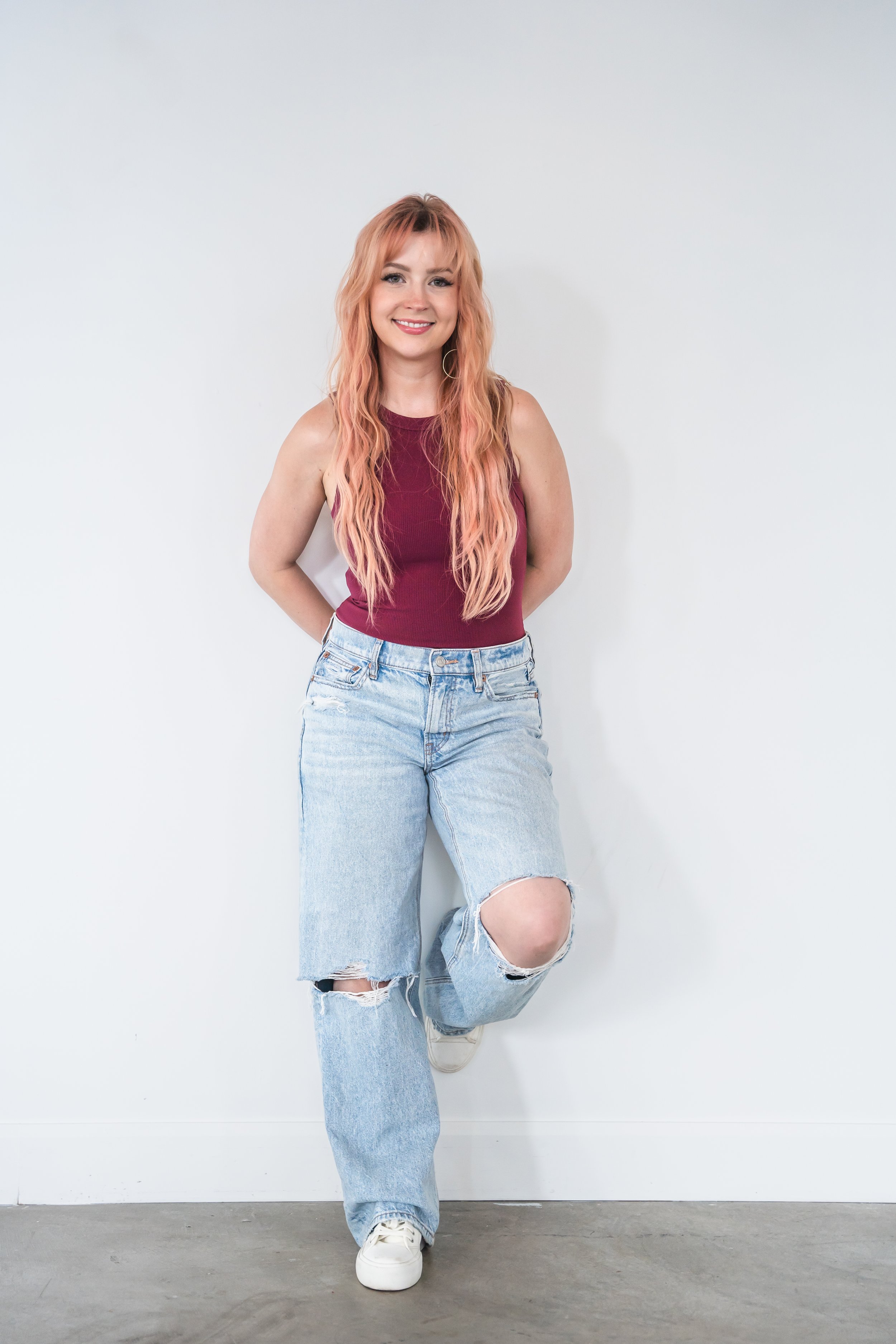 A hair stylist from Brantford Ontario Canada posing in front of a white wall, her one foot is up against the wall behind her, her hands are behind her back. She's smiling softly at the camera.