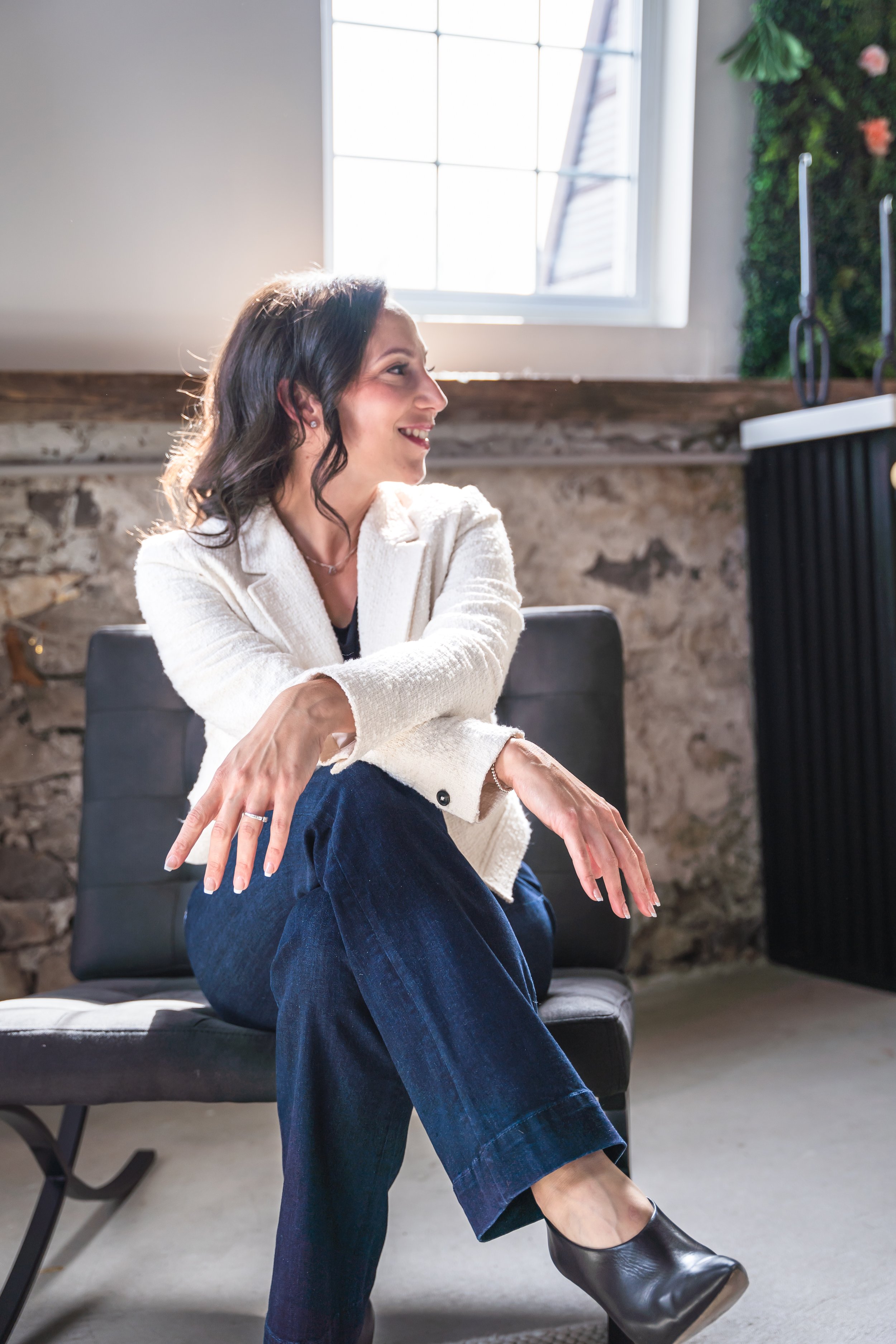 An event planner from Brantford Ontario Canada sitting on a chair in front of a window and a brick wall. Her legs are crossed at her knee and her arms are rested on her legs. She's smiling while looking off to the side.