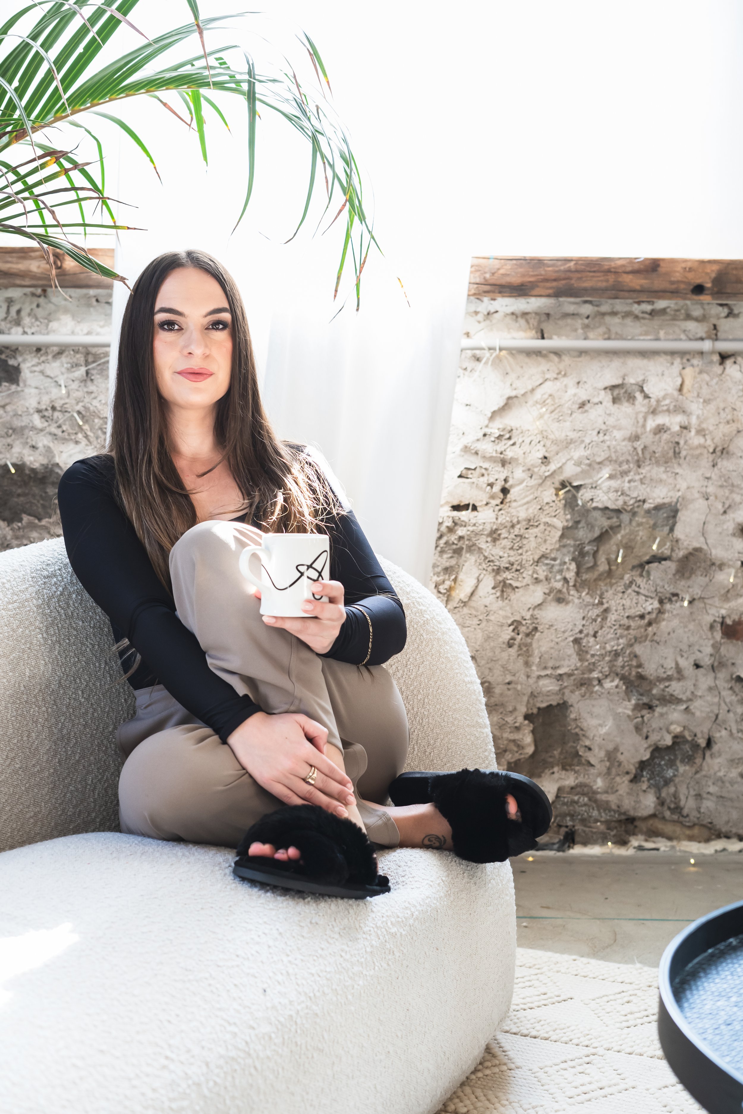 A consultant from Brantford Ontario Canada sitting cross legged on a cream couch with a cup of coffee in her hand. She's looking directly at the camera with a passive look on her face.