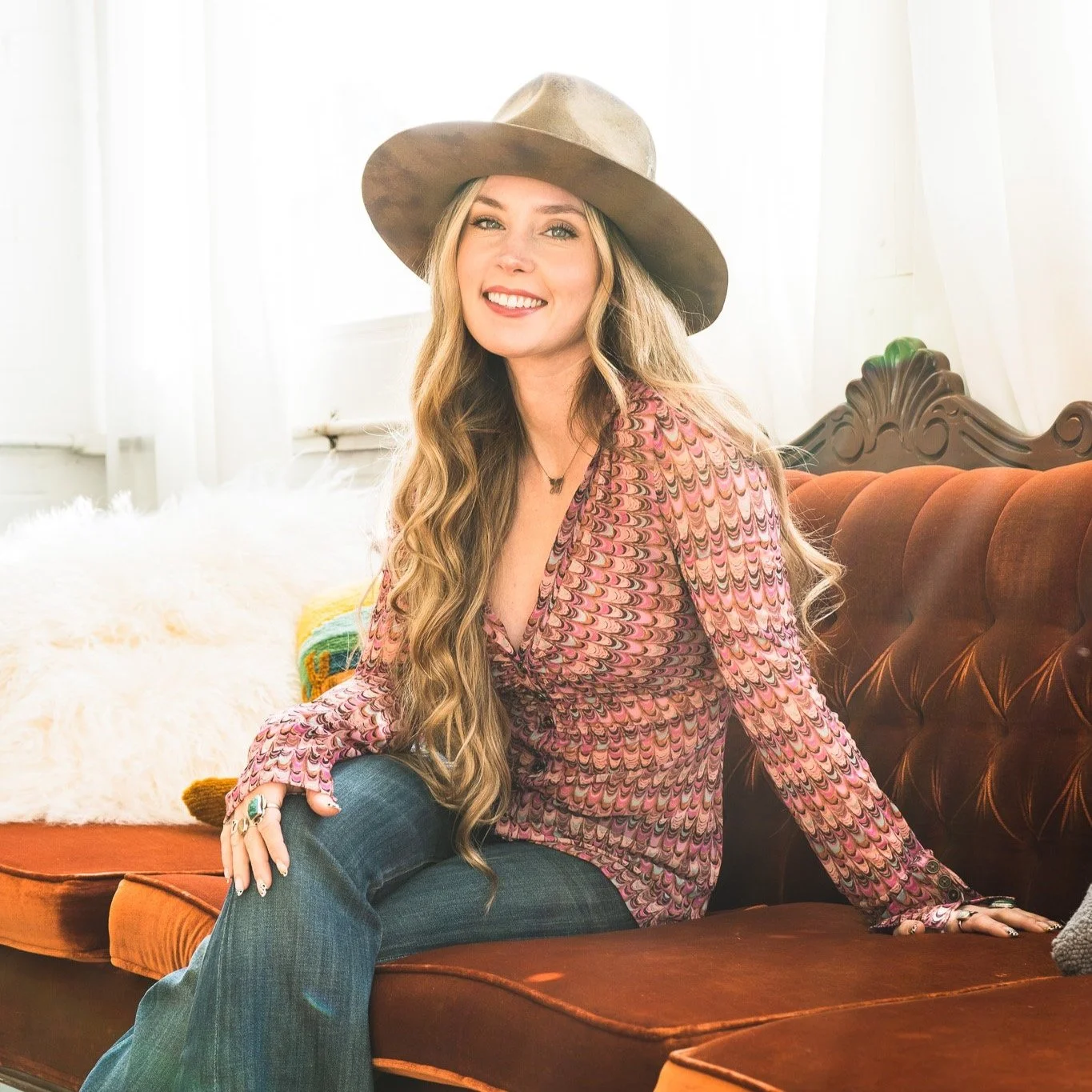 A Brand photographer's view of woman sitting on a vintage orange velvet couch, wearing a wide-brimmed hat and a colorful patterned blouse, smiling at the camera in a well-lit room with white walls and a white fluffy blanket.