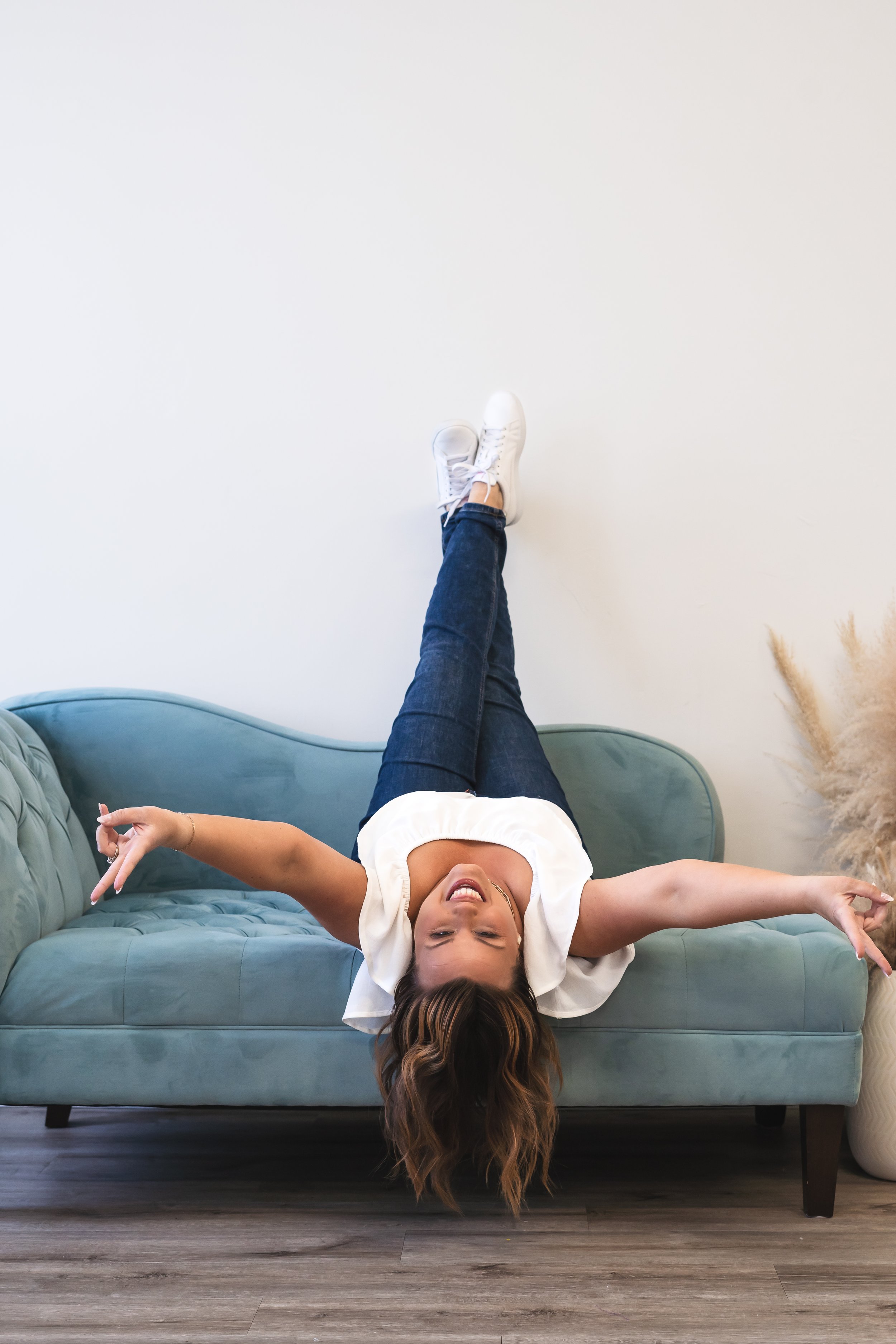A content creator from London Ontario Canada sitting upside down on a teal couch with her ankles crossed up in the air and making the peace sign with both hands. She's smiling at the camera.
