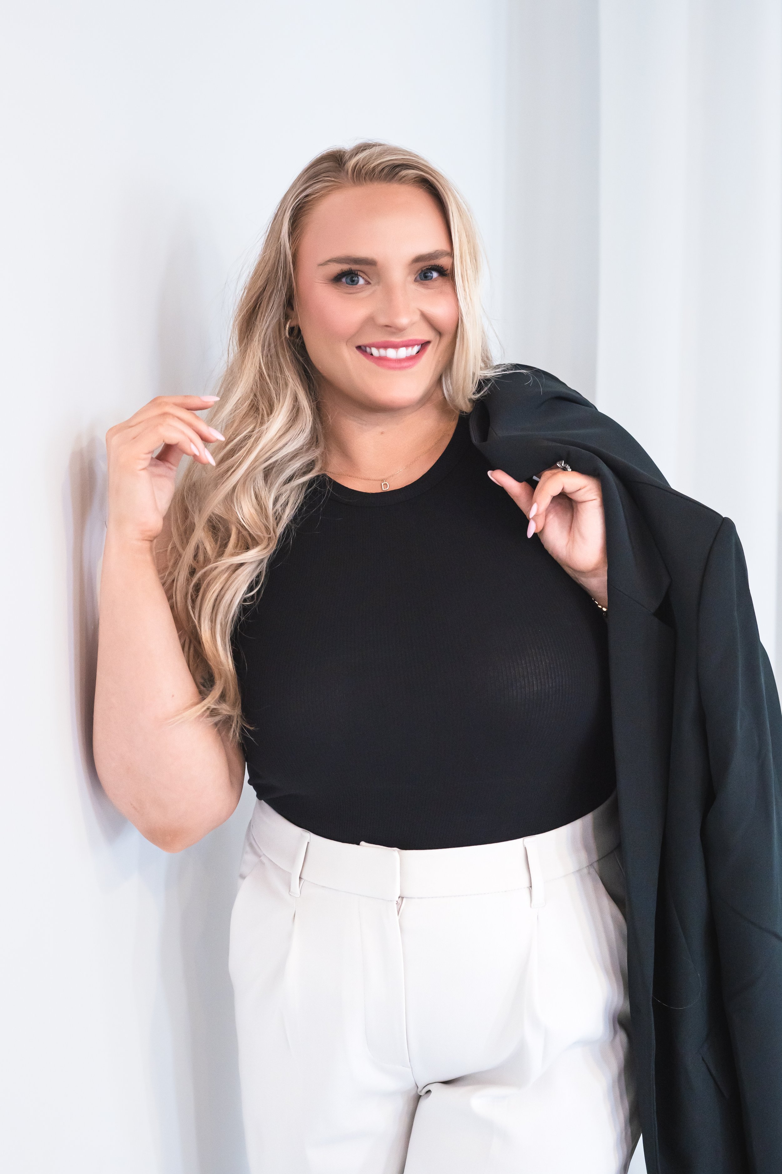 A realtor from Hamilton Ontario Canada leaning up against a white wall. Playing with her hair in one hand and holding a blazer over her shoulder with the other.