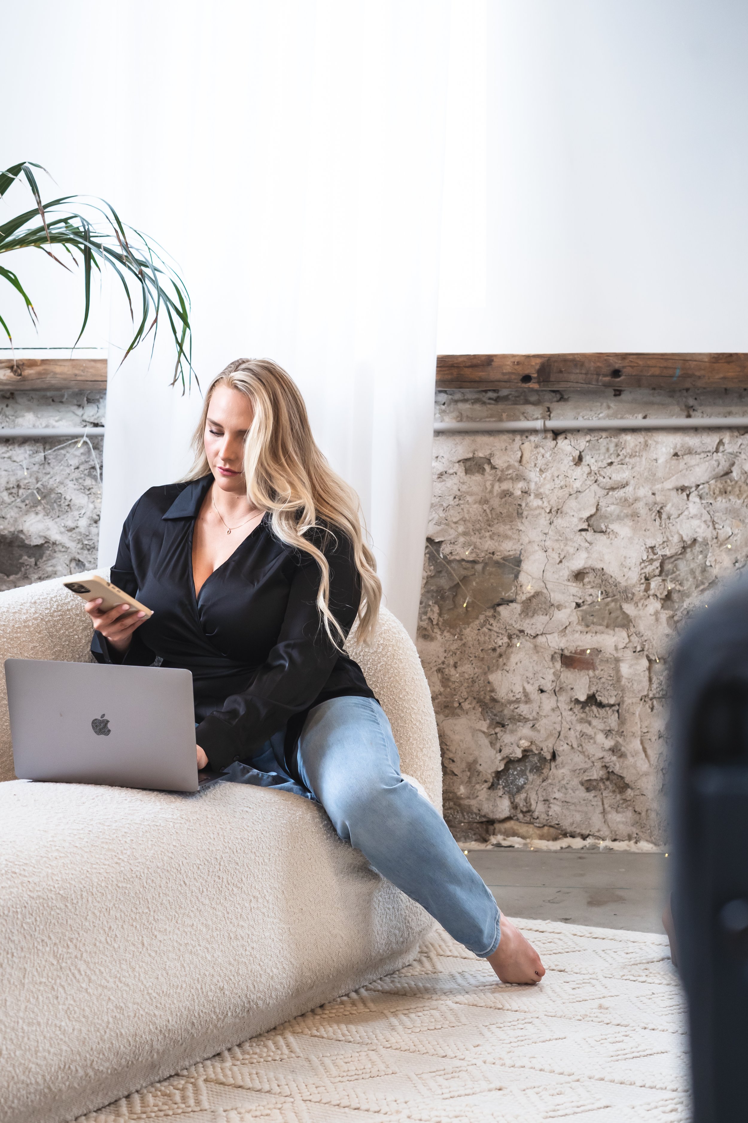A real estate agent from Hamilton Ontario Canada posed on a cream couch with her phone in her hand and her MacBook open in front of her.