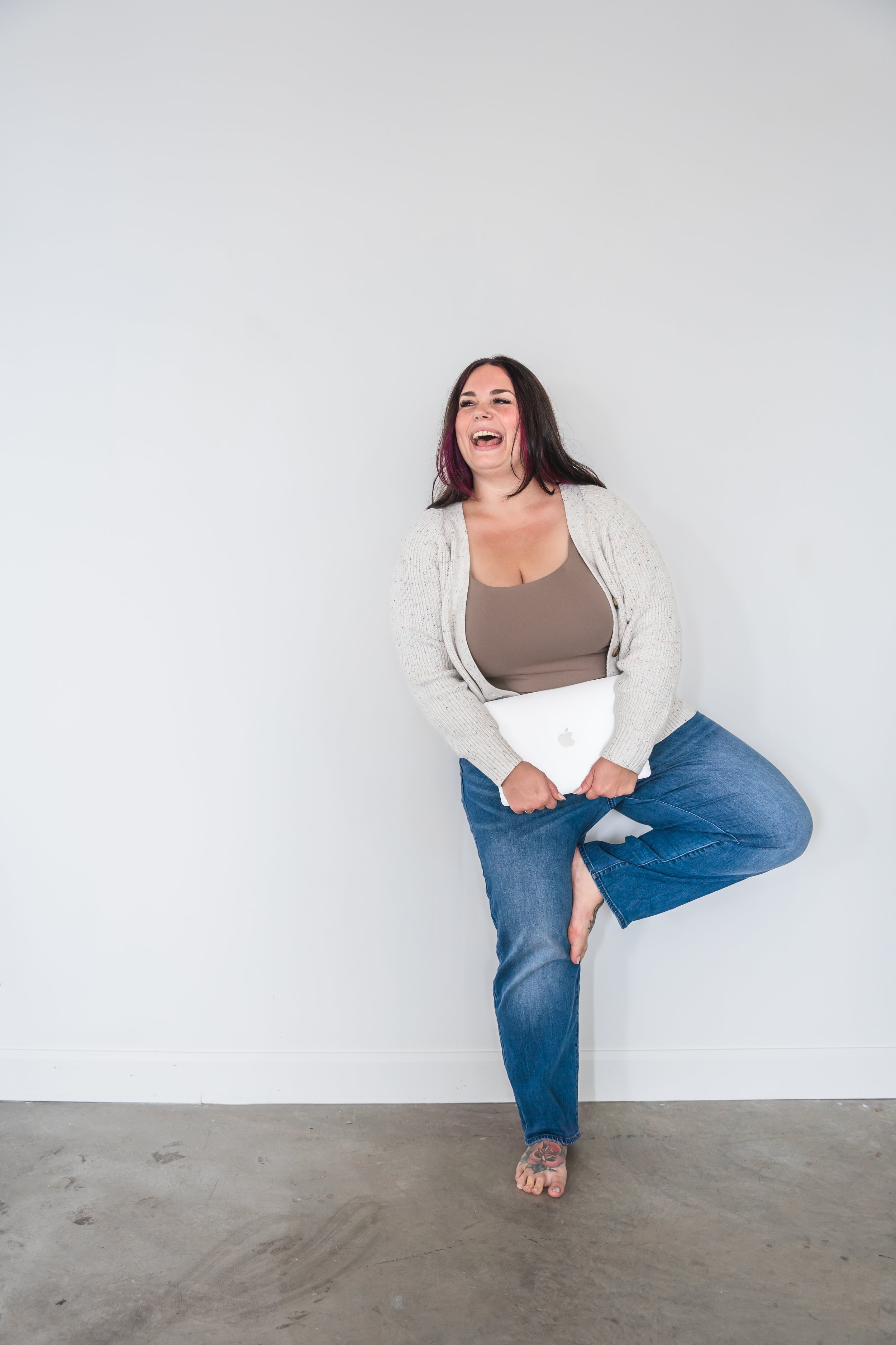 A therapist from Brantford Ontario Canada posing in front of a white wall with one let up at her knee, holding her laptop in front of her and laughing into the camera.