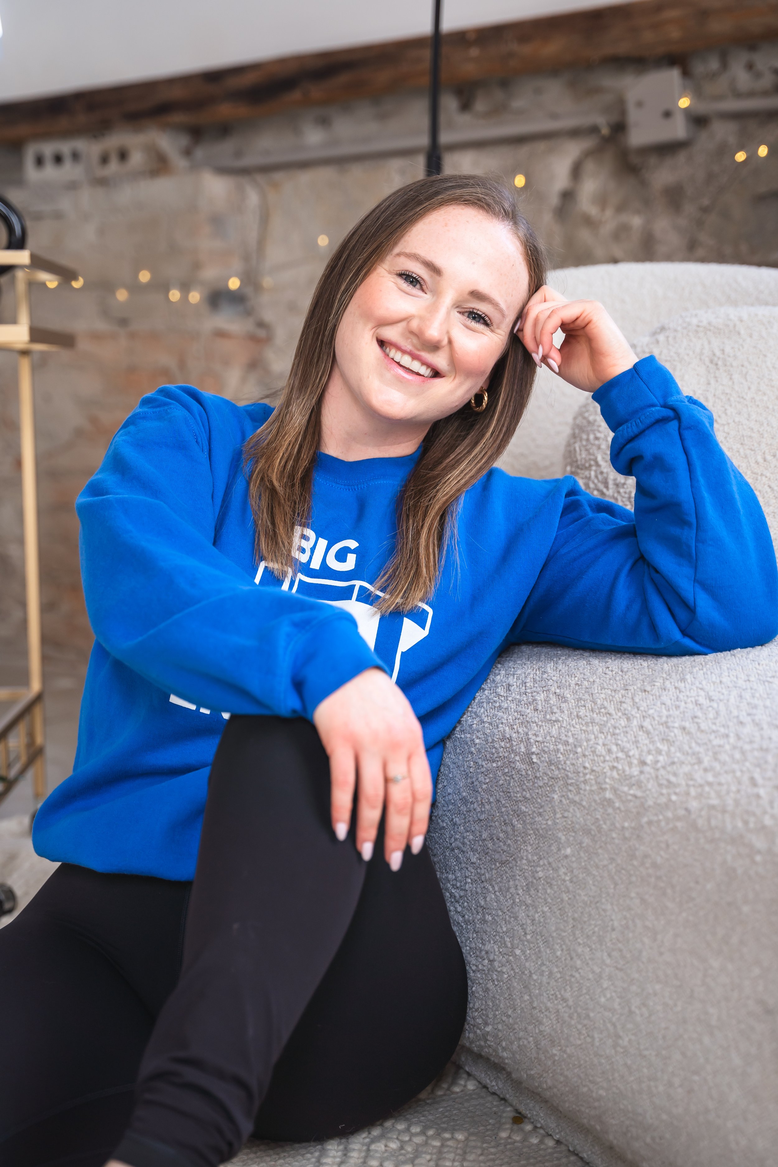 A personal fitness trainer from Brantford Ontario Canada sitting next to a cream couch, leaning one arm up with her hand resting on her hand.