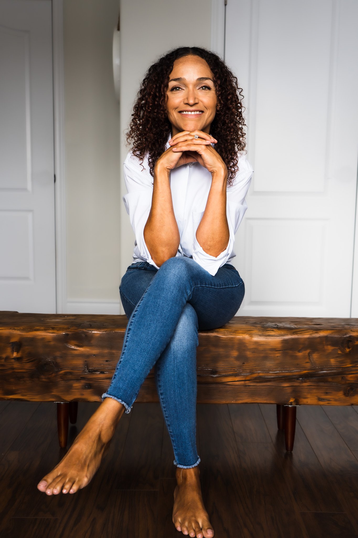 A wellness consultant in London Ontario posing for a brand photographer on a wooden bench, legs crossed with her elbows on her knees, chin resting on her hands.