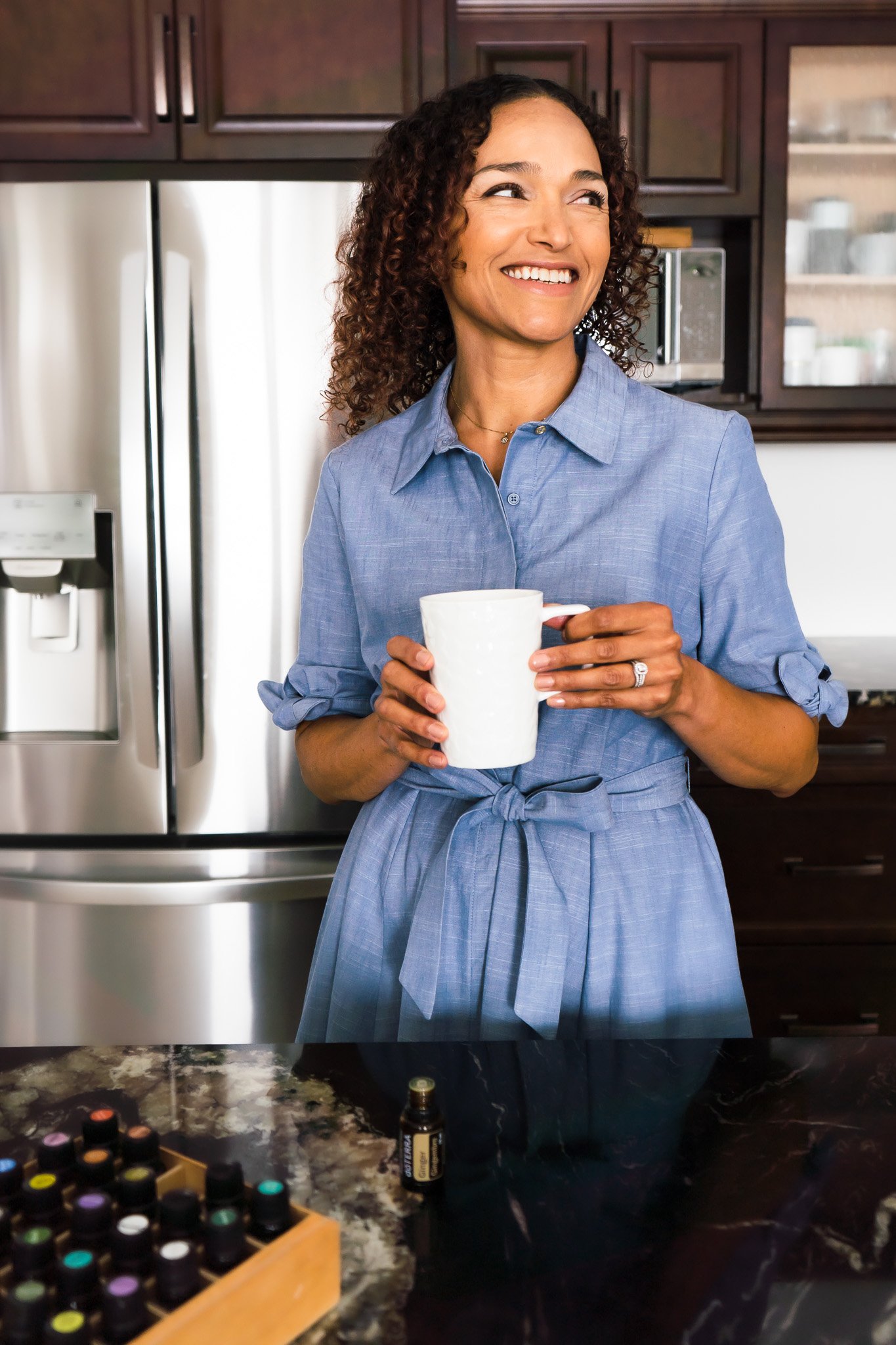 A wellness consultant posing in her kitchen holding a cup of tea, she's smiling into the distance, her collection of essential oils in the foreground.