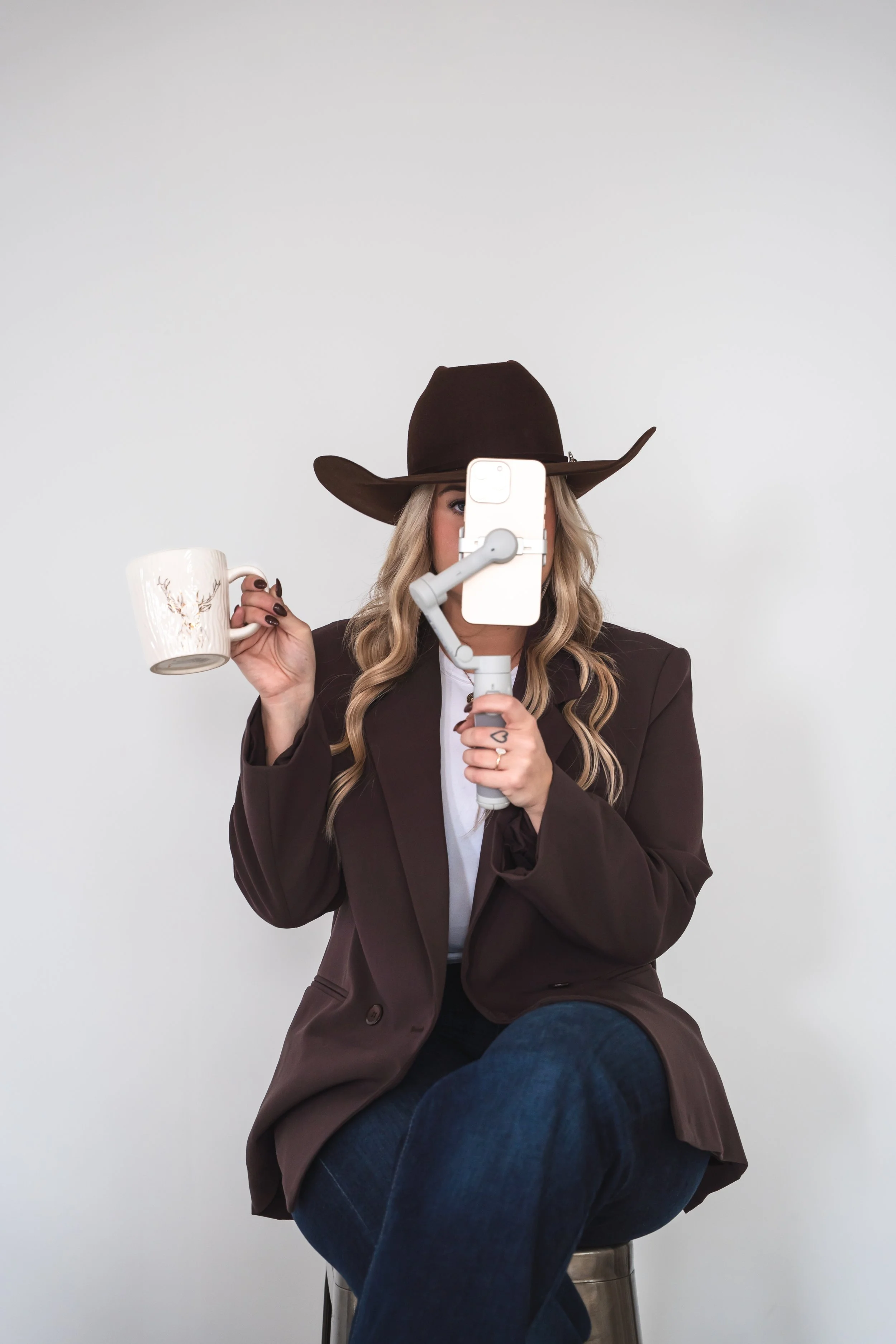a brand photographers view of a content creator holding her phone up in front of her face, a coffee mug in her hand wearing a cowboy hat.