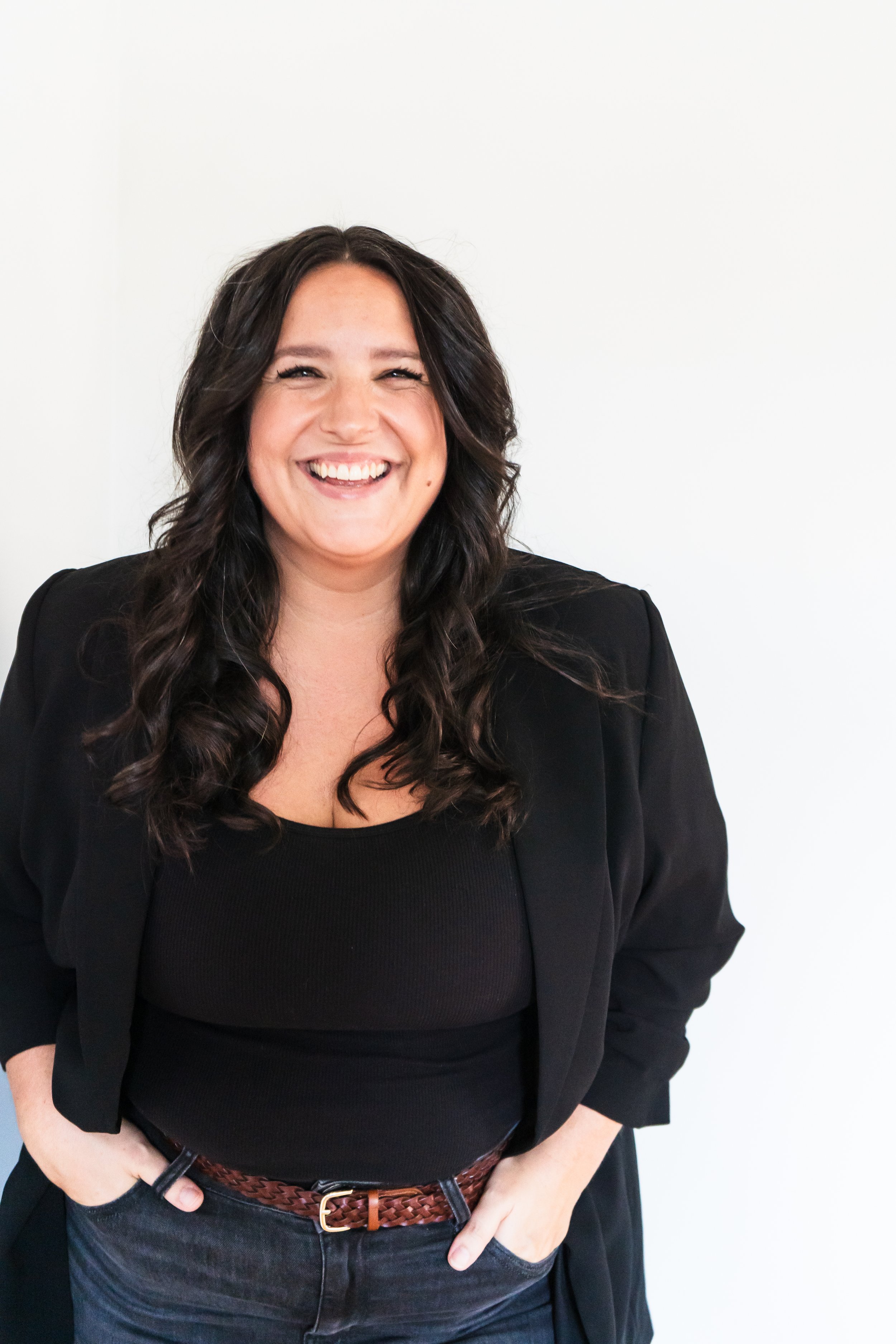 A copywriter from Kitchener Ontario Canada posing in front of a white wall with her hands in her pockets, she's smiling confidently into the camera.