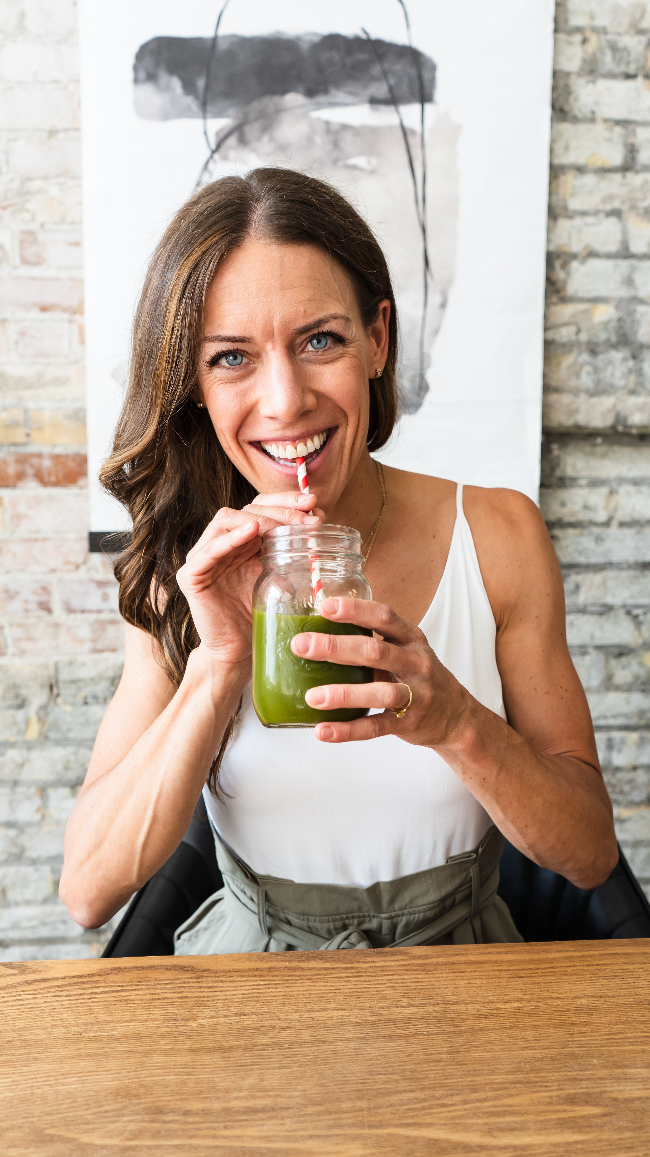 A naturopathic doctor from Kitchener Ontario Canada sitting at a wooden desk with a brick wall in the background. She's drinking a green smoothie from a mason jar while smiling at the camera.