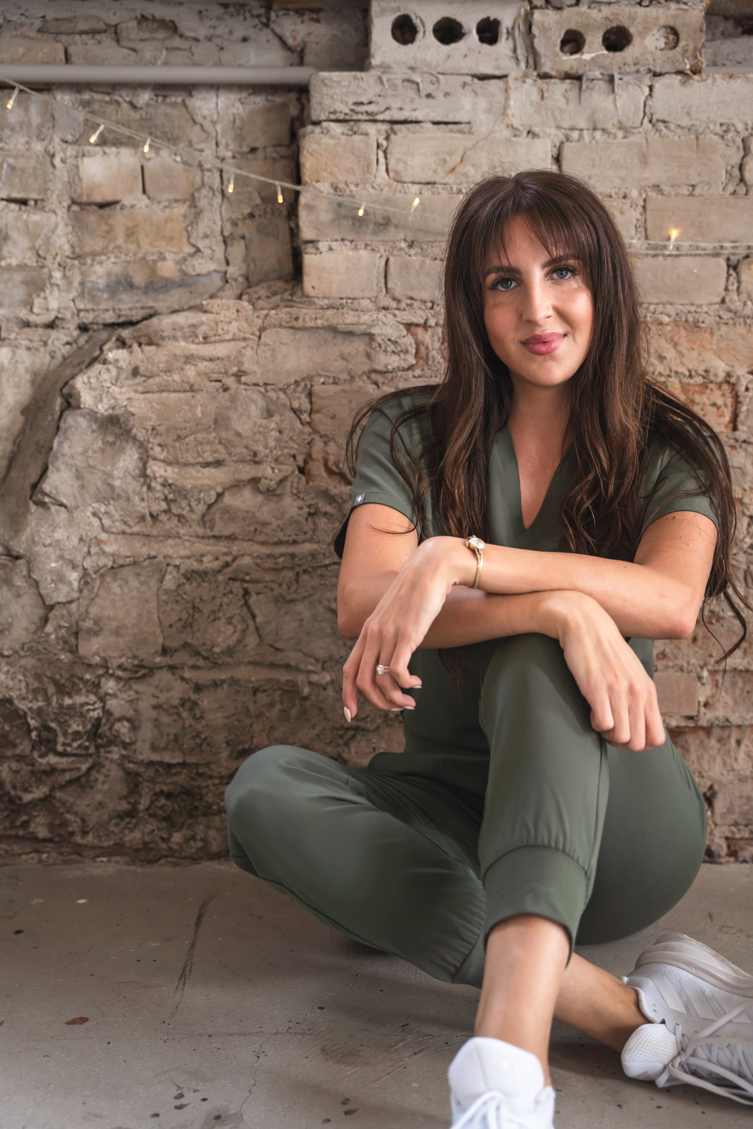 A medical aesthetician from Brantford Ontario Canada posing in front of a brick wall wearing stylish green scrubs. She's sitting with her legs slightly crossed and her hands draped across her knee.