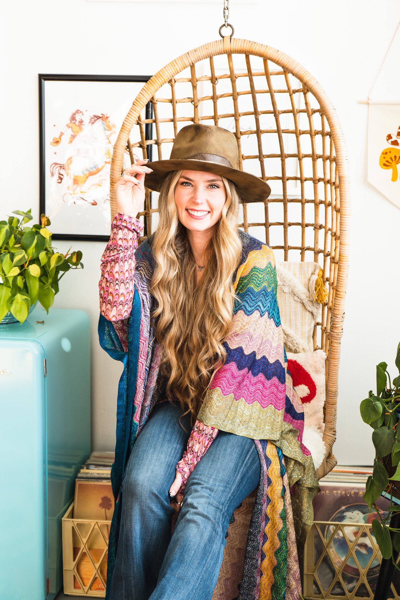 A creative business owner from Cambridge Ontario Canada sitting in a hanging wicker chair in her workshop, a teal blue fridge beside her and artwork in the background.
