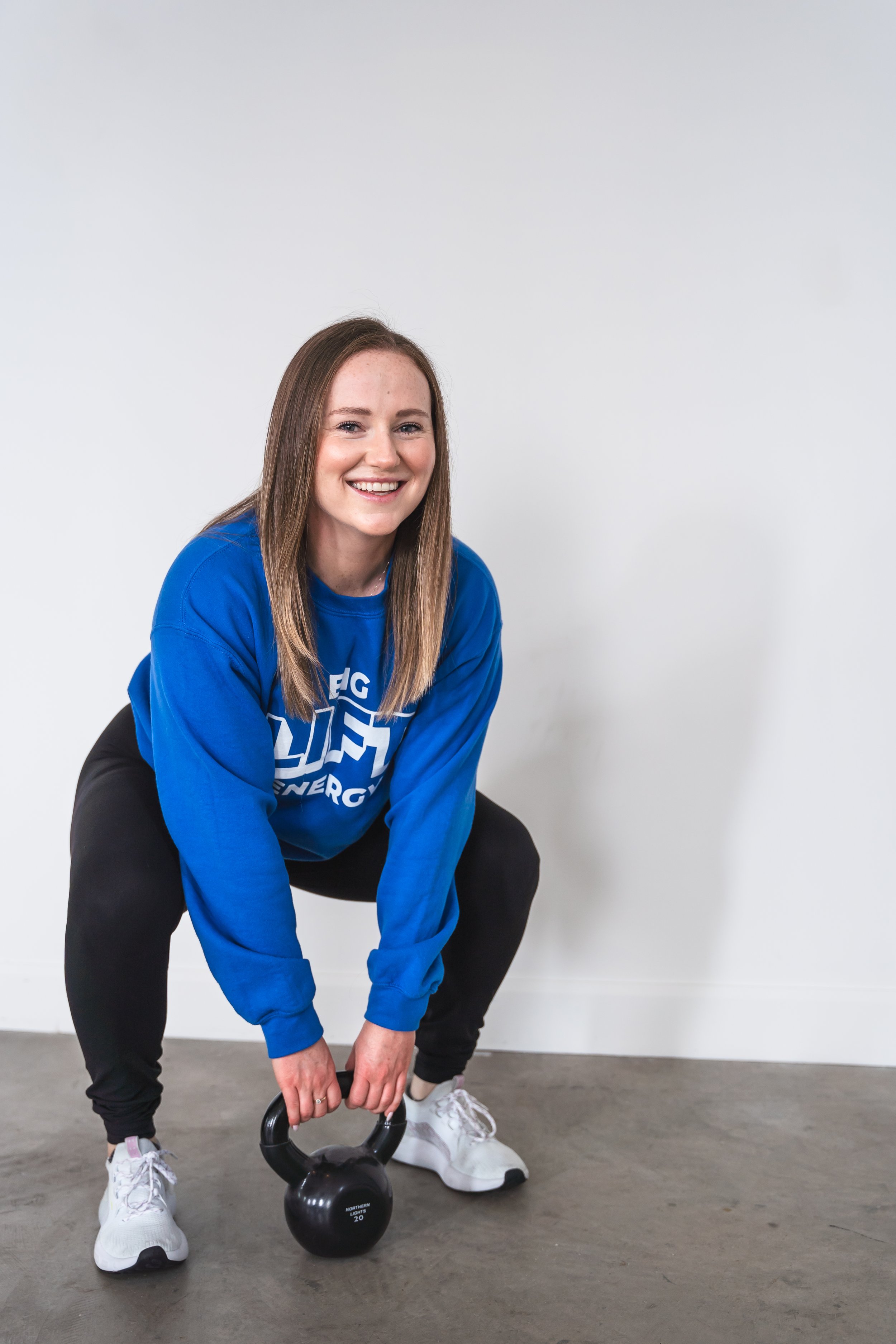 A fitness coach from Brantford Ontario Canada demonstrating a squat with a kettle bell in front of a white wall while smiling at the camera.