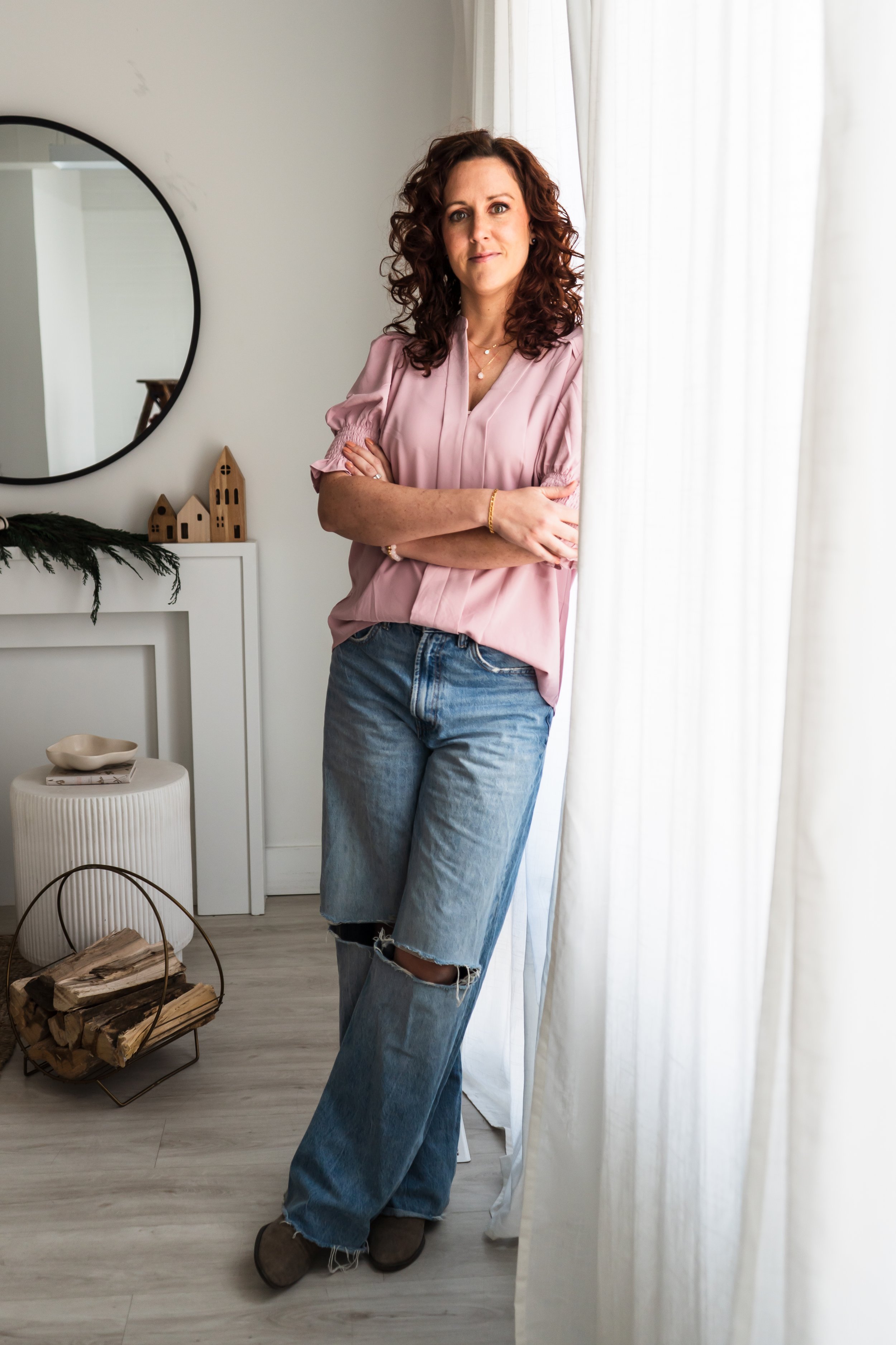 A jewelry designer from Burlington Ontario Canada leaning up against a bright window with white curtains. A mirror is on the wall behind her.