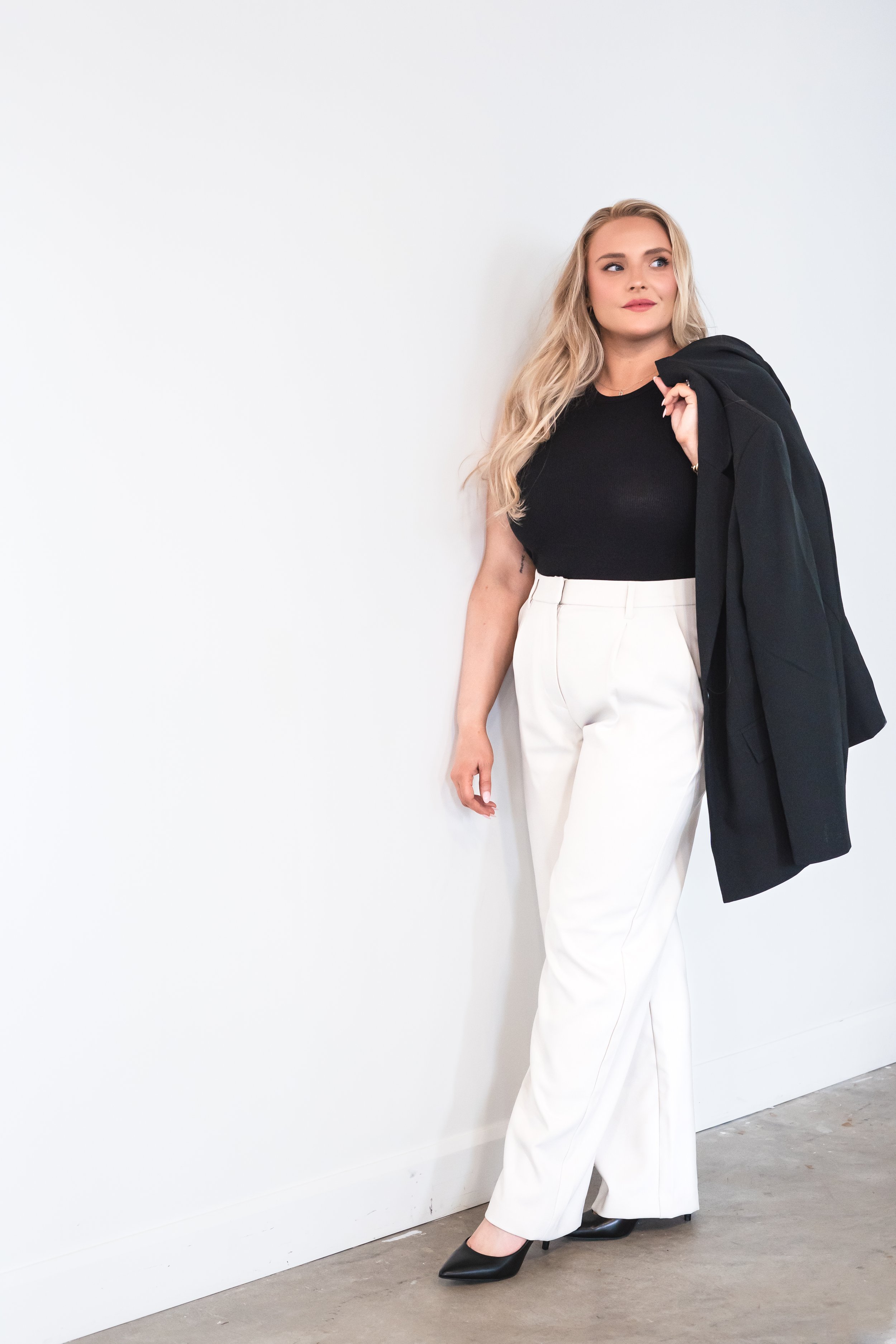 A Canadian brand photographer's view of a Hamilton Ontario realtor beside a white wall with light pants and a black shirt, a blazer over her shoulder.