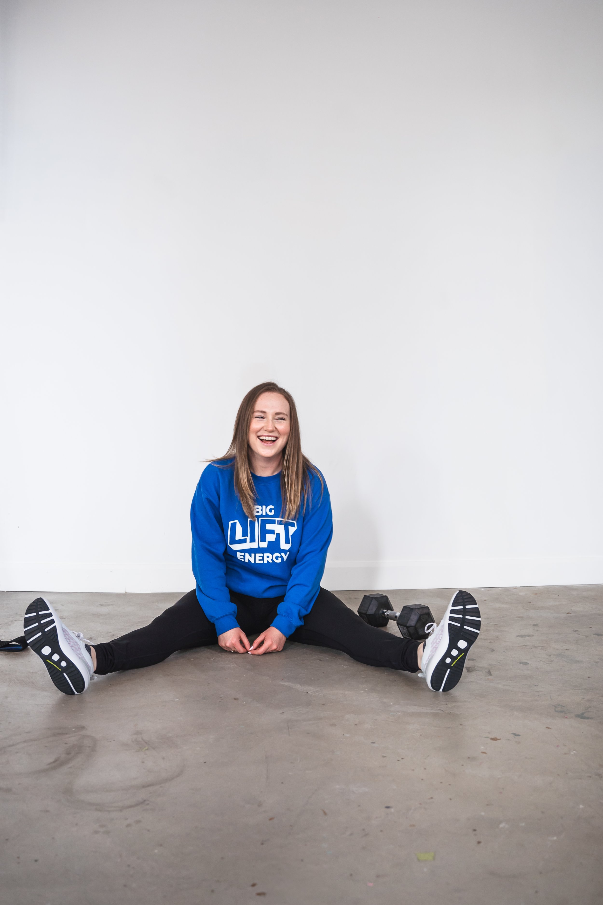 A personal fitness trainer from Brantford Ontario Canada sitting on the floor in front of a white wall with her legs spread out in front of her. She's laughing at the camera.