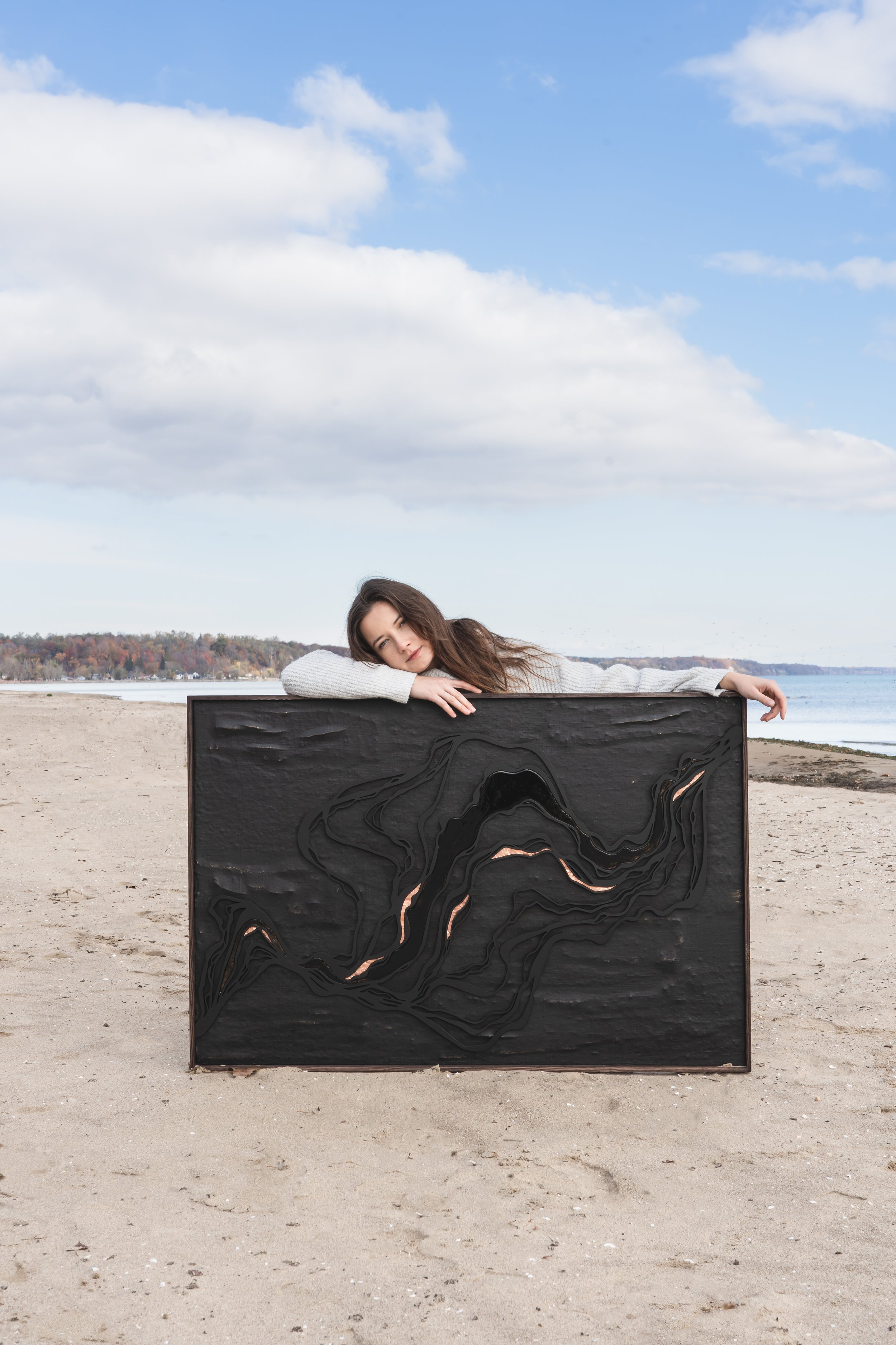 An artist from Brantford Ontario Canada posing with her art on a beach with a blue sky. She's behind her art and leaning across the top of it.