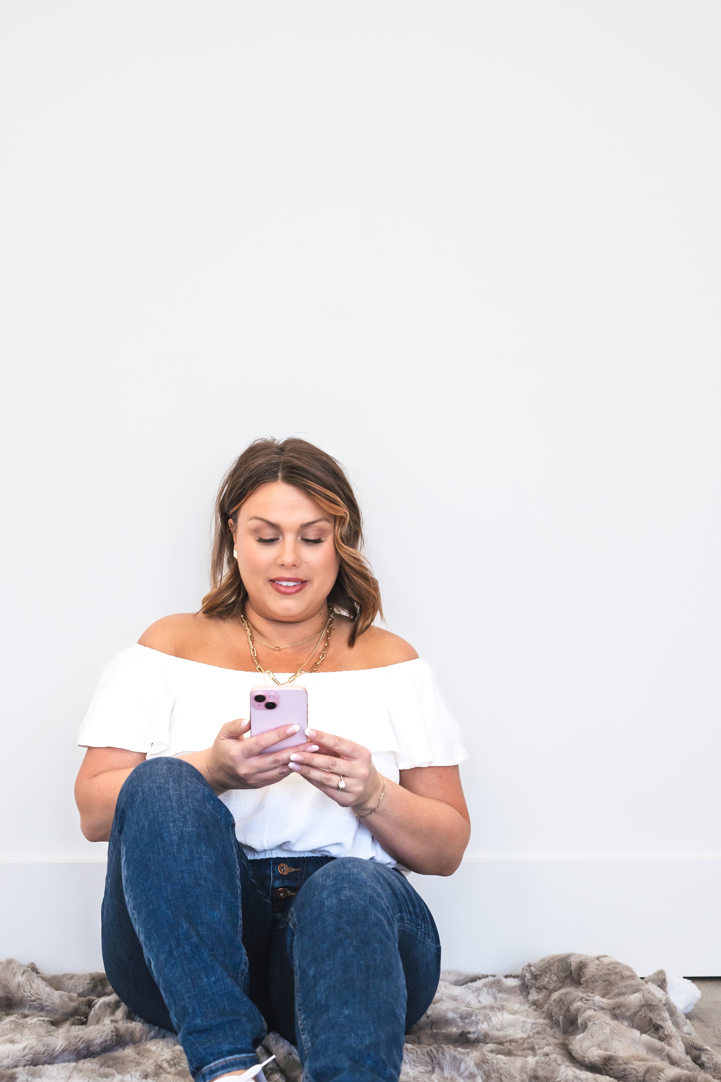 A content creator from London Ontario Canada sitting on the floor in front of a white wall with one leg up. She's looking down at her phone.