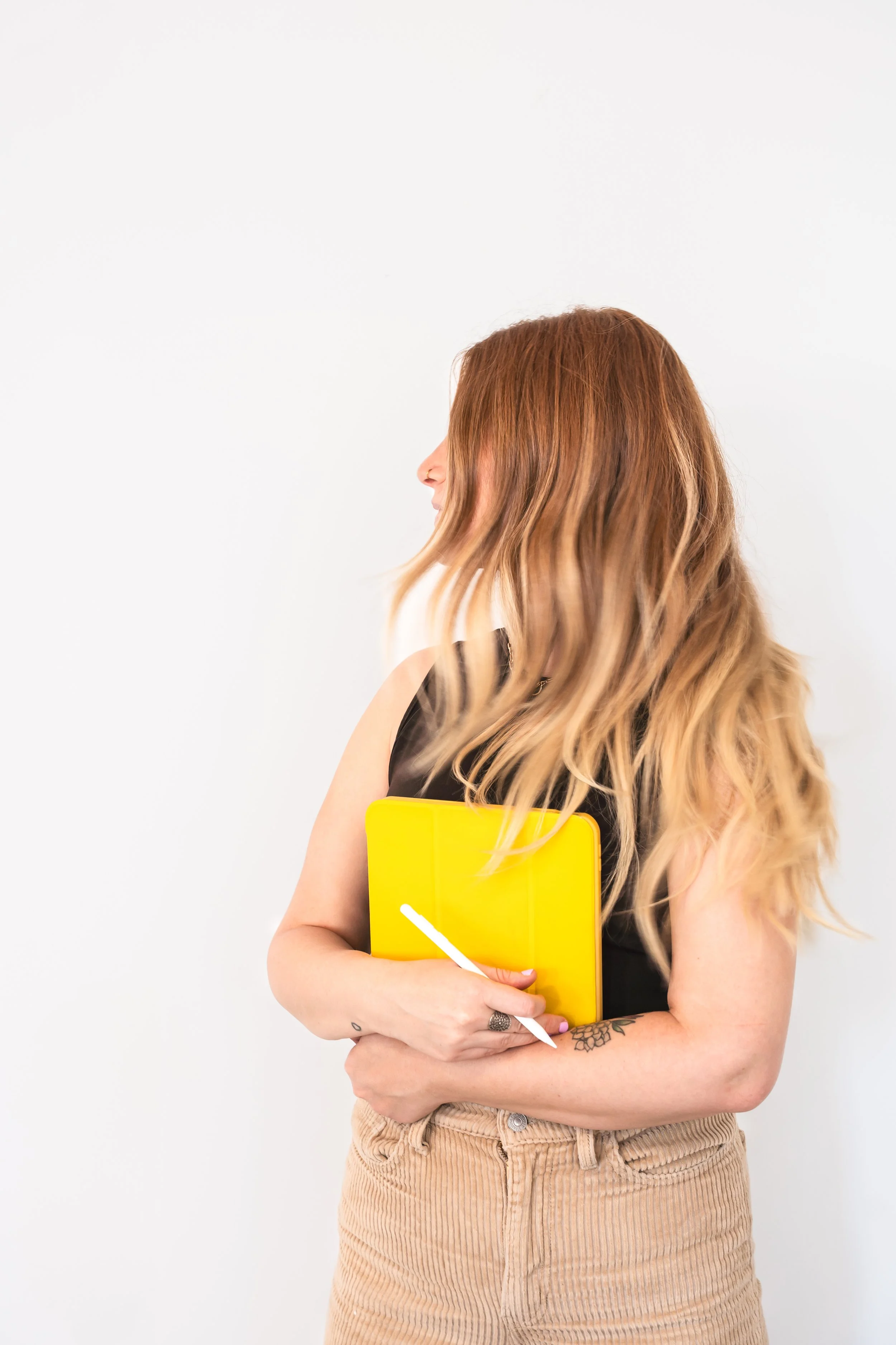 A Canadian brand photographers view of a graphic designer in front of a white wall holding her iPad in a yellow case while turning her head.