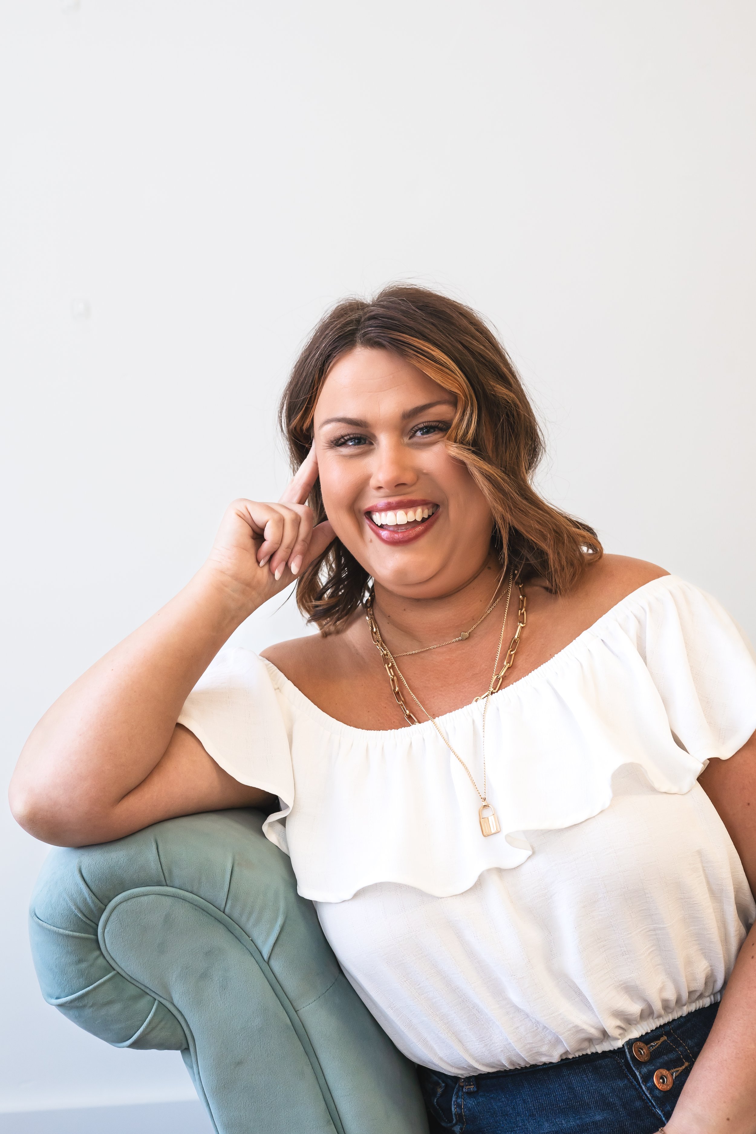 A content creator sitting on a blue couch in front of a white wall. She's got her elbow up on the side and leaning on her hand, smiling confidently at the camera.