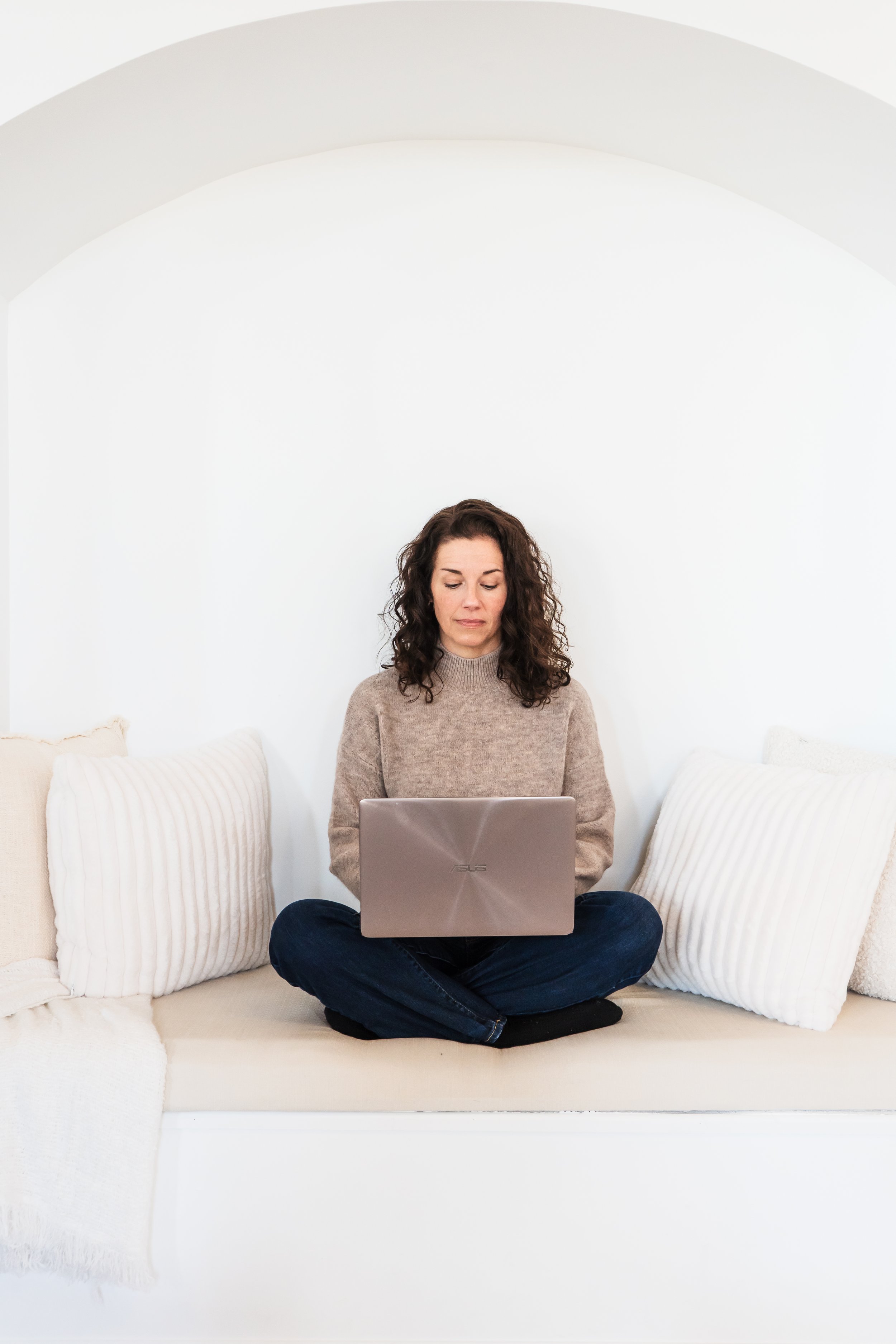 A therapist from Kitchener Ontario Canada sitting cross legged on a white bench with her MacBook laptop in her lap, she's looking down at her laptop.