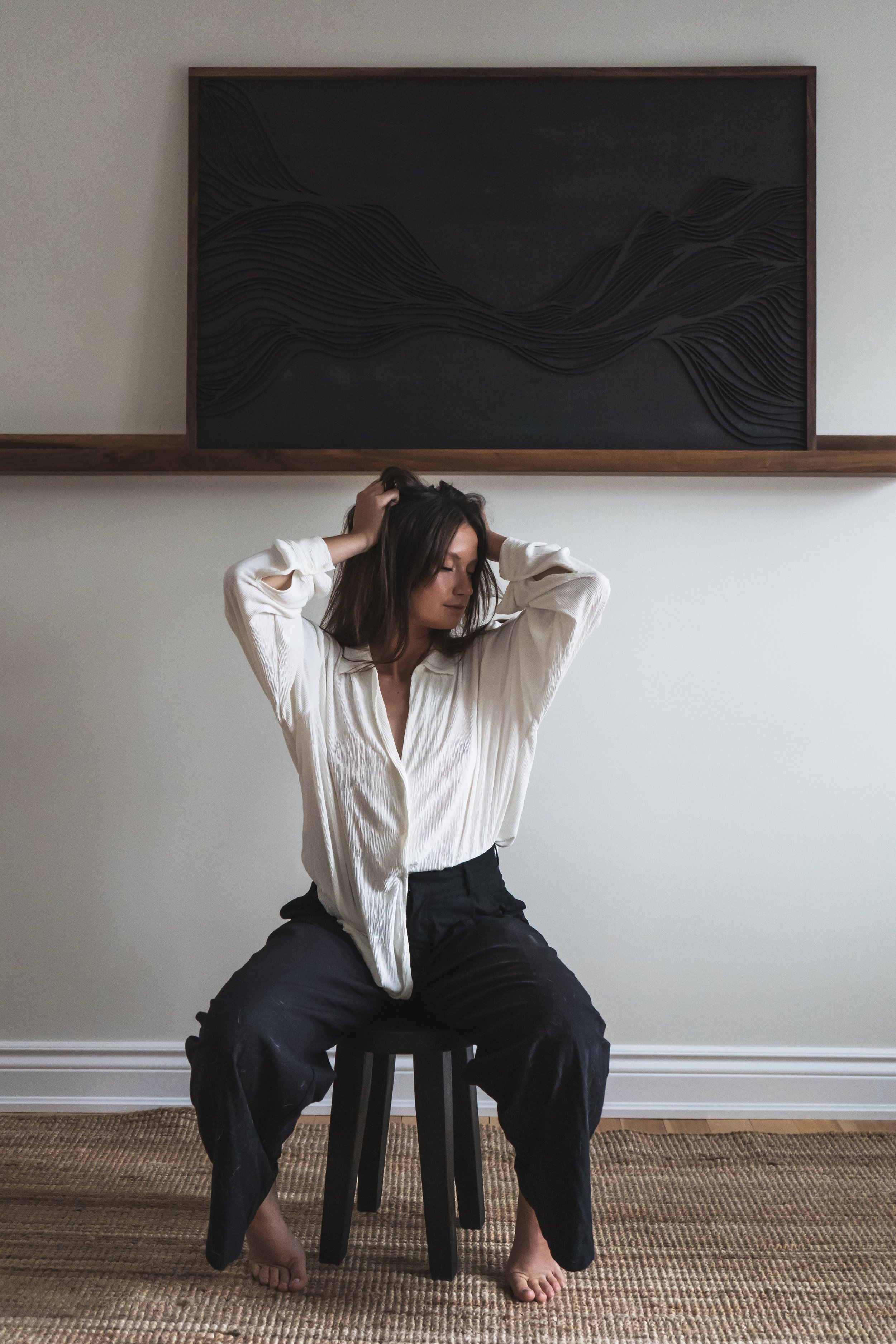 An artist from Brantford Ontario Canada sitting on a stool, her art on a shelf on the wall be hind her. She's got her hands running through her hair.