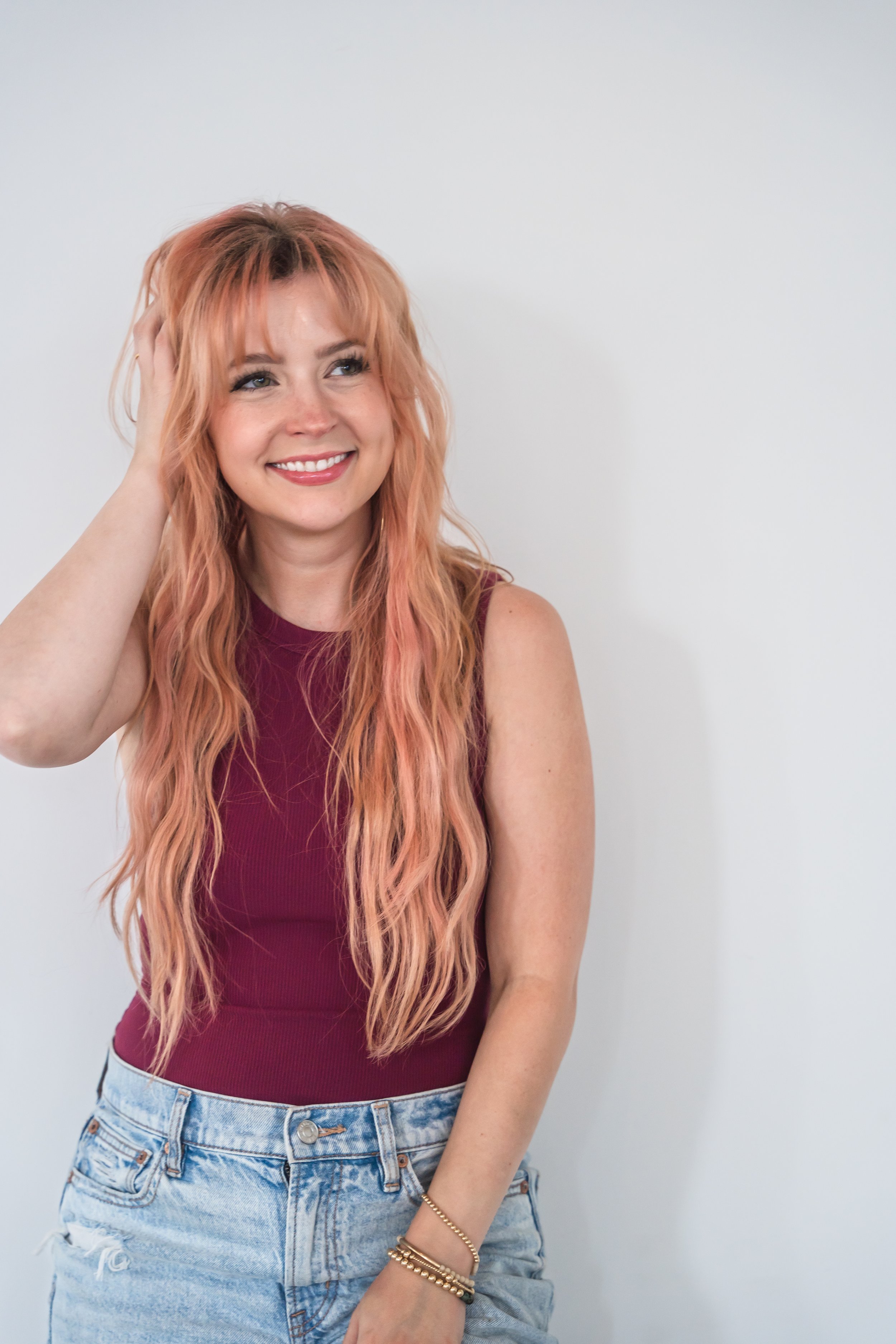 A hair stylist from Brantford Ontario Canada posing in front of a white wall, she's got one hand bunching up her light pink hair and smiling just past the camera.