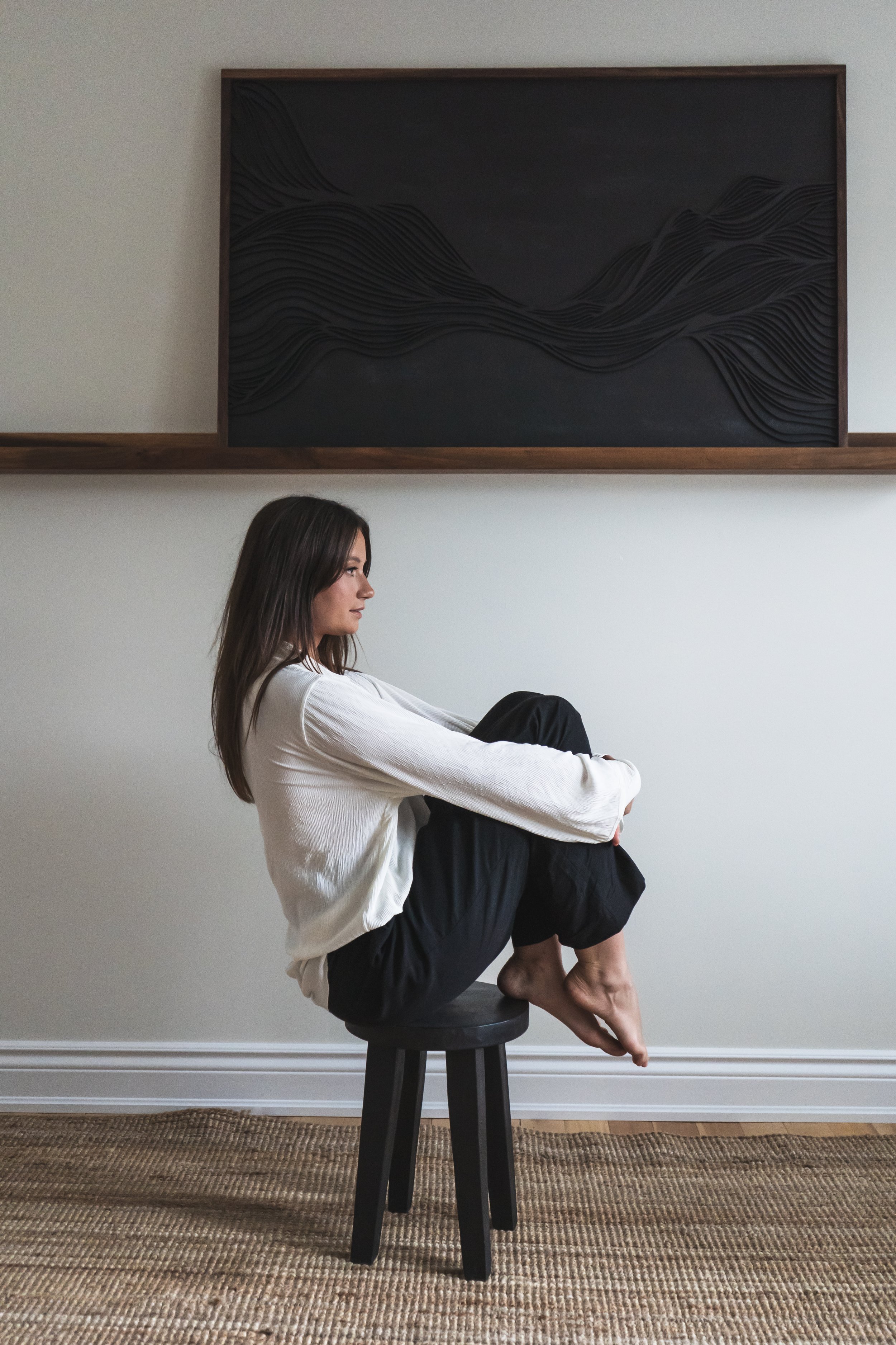 An artist from Brantford Ontario Canada posing on a stool in front of her art that's on a shelf on the wall behind her.