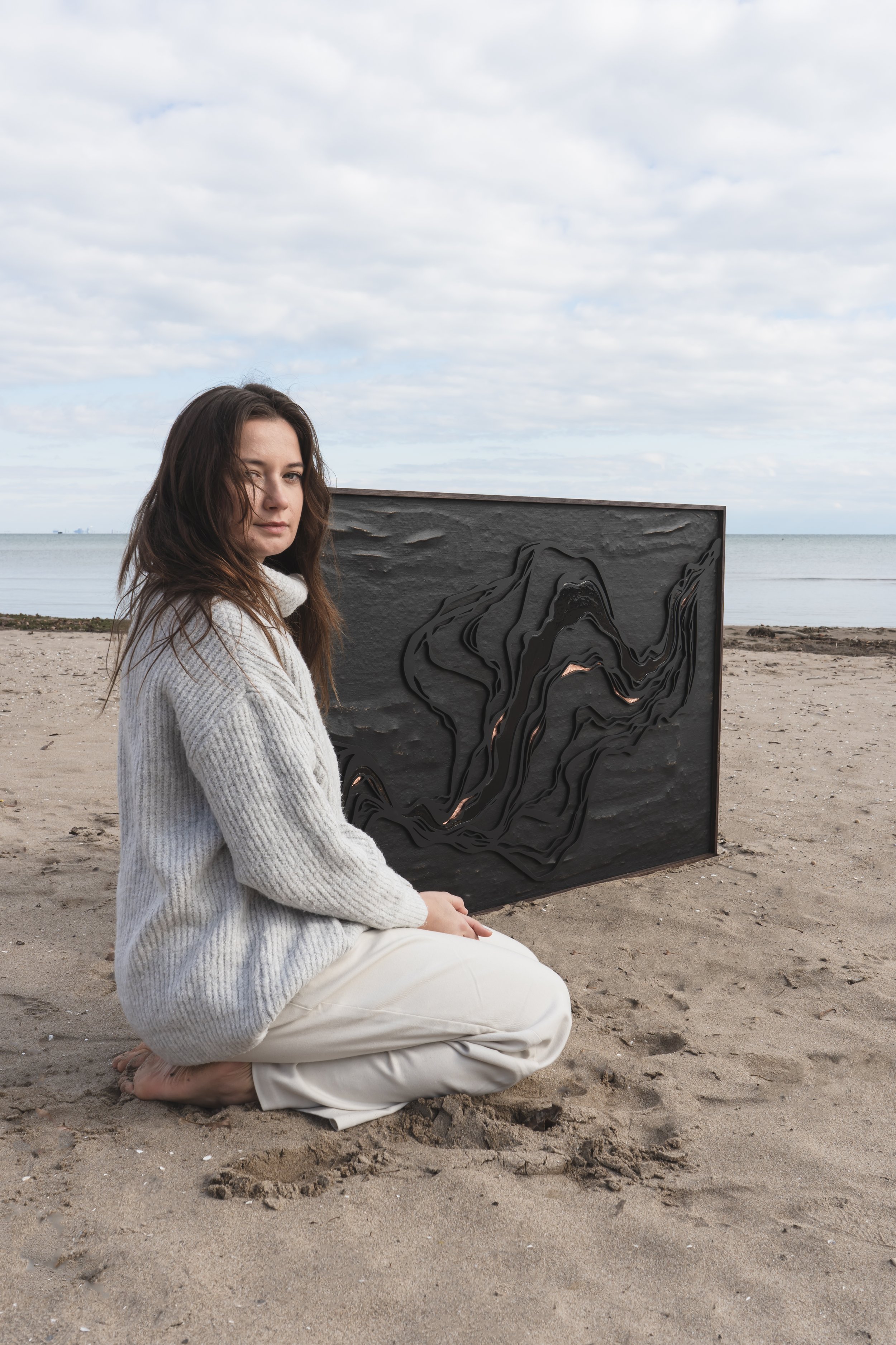 An artist from Brantford Ontario Canada kneeling in the sand in front of her art with a blue sky behind her. She's looking softly at the camera.