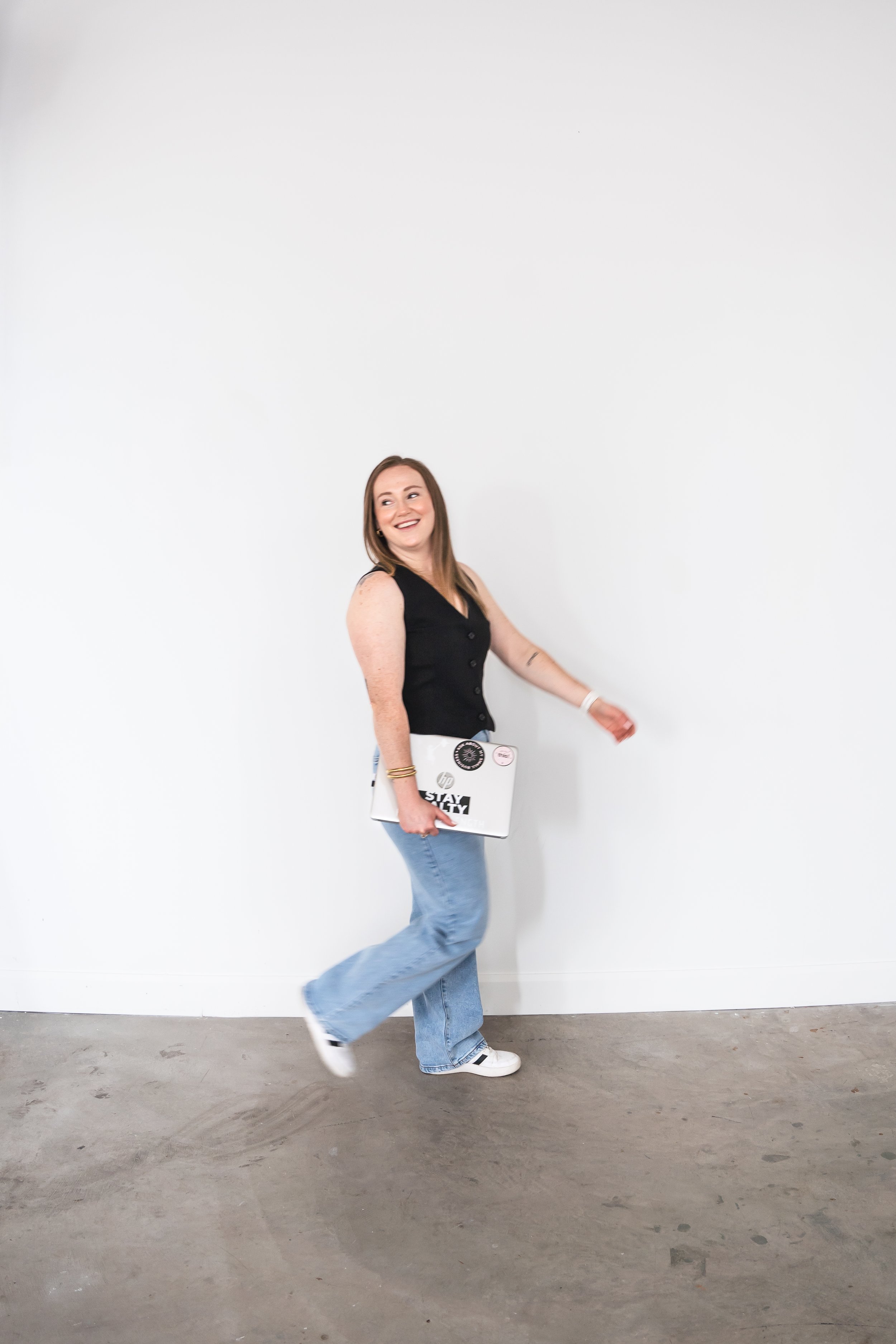 A fitness coach from Brantford Ontario Canada walking across a white wall, her laptop in her hand at her side. She's smiling back over her shoulder.