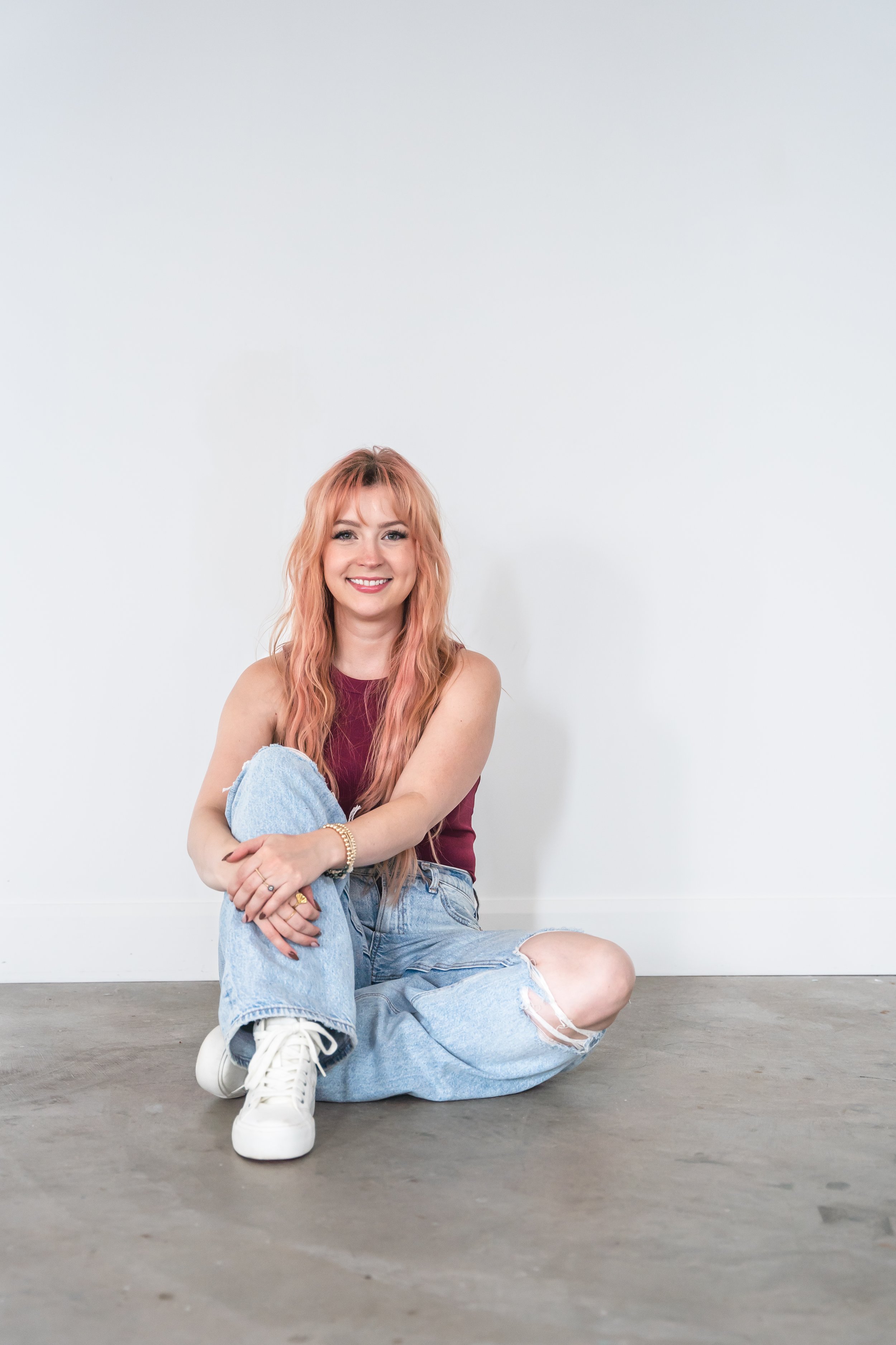 A hair stylist from Brantford Ontario Canada sitting in front of a white wall with one leg up and her hands crossed in front of her knee. She's smiling confidently at the camera.